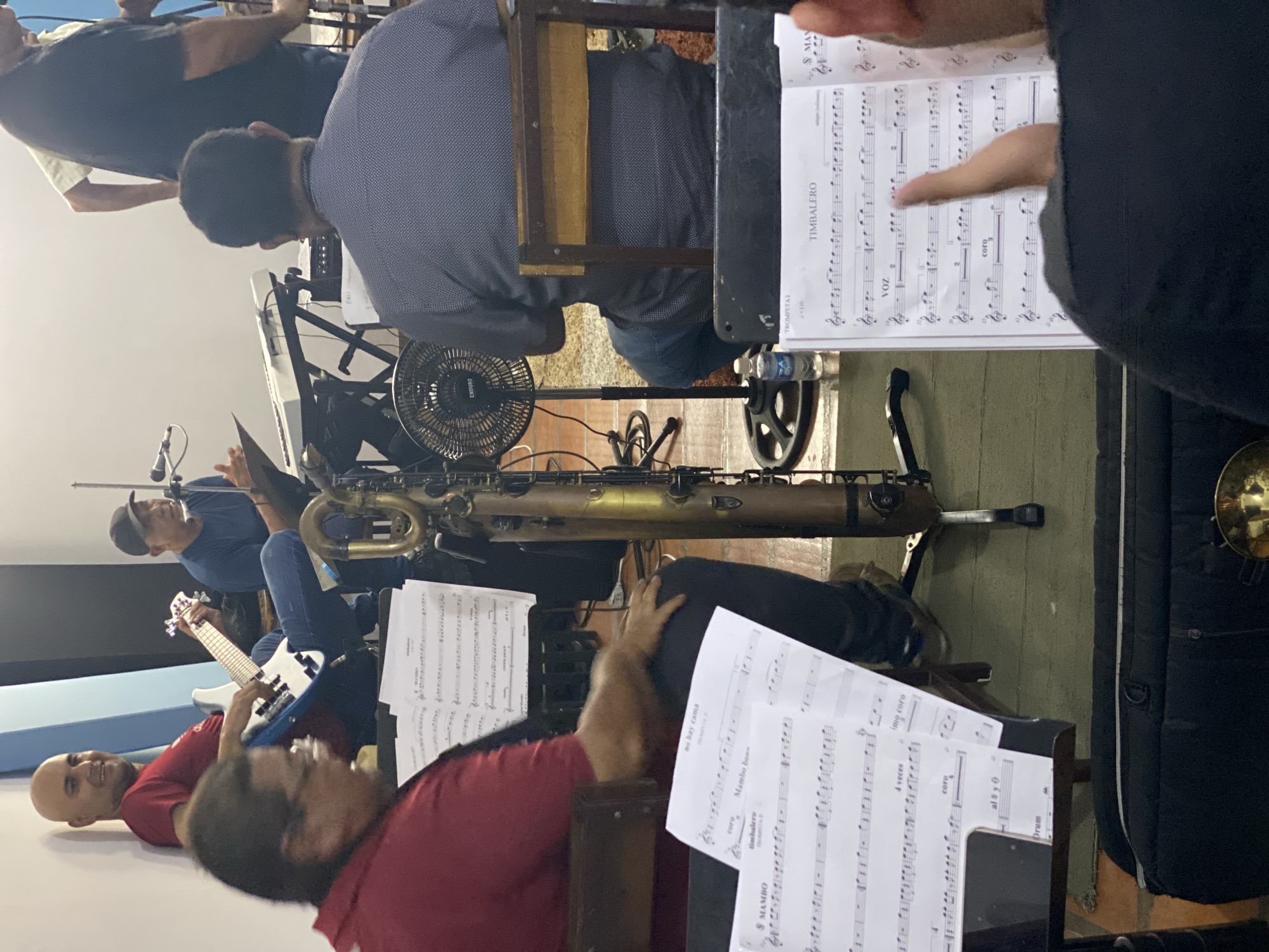 The image depicts a group of musicians rehearsing indoors, presumably in Tinaquillo, Venezuela given the text on the sheet music. Several men are visible, playing various instruments including a bass guitar and a saxophone. Sheet music is placed on stands in front of them. A fan is placed near the musicians. The lighting is somewhat dim. The overall setting appears to be a rehearsal space.