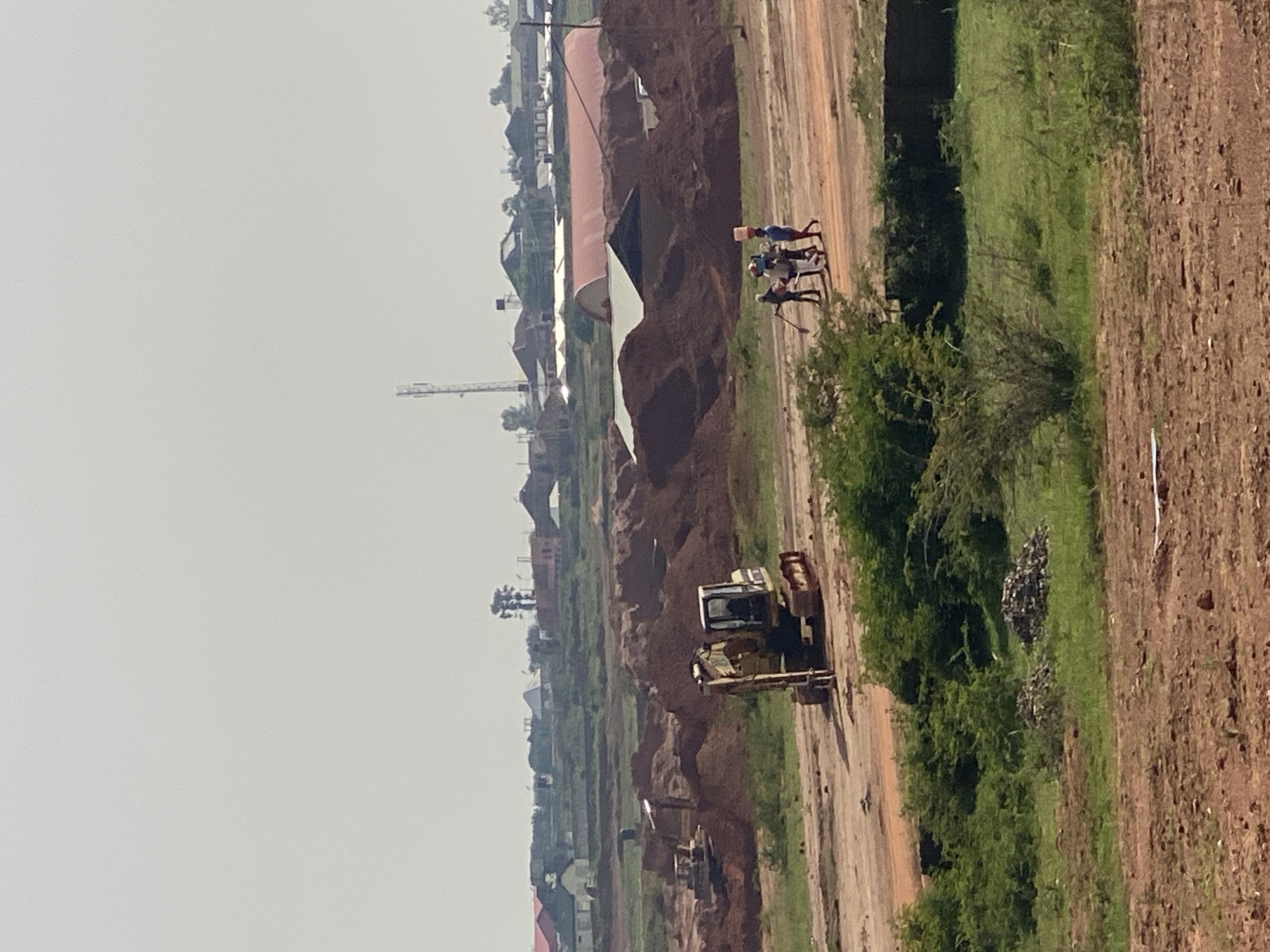The image depicts an outdoor construction or excavation site, likely in Jos, Nigeria, under a hazy sky. In the foreground, reddish-brown earth is visible, with some patches of green vegetation. A dirt road or path runs through the mid-ground, leading to a larger construction area.
Dominating the mid-ground is a yellow excavator, parked on the dirt path. Further behind and to the left, another construction vehicle, possibly a bulldozer, is partially visible amongst piles of reddish-brown earth. Large mounds of earth, suggesting excavation or material staging, are scattered throughout the scene.
In the background, a cluster of buildings with various roof types, including what appears to be a large, arched red-roofed structure, are visible. A tall communication tower stands in the distance.
A group of four individuals is walking on the dirt path in the mid-ground, moving from left to right. One person in the center appears to be carrying a large, round object on their head. The individuals are dressed in casual clothing, and their interaction appears to be that of a group walking together. The overall impression is that of ongoing work at a large-scale construction or earthmoving operation.