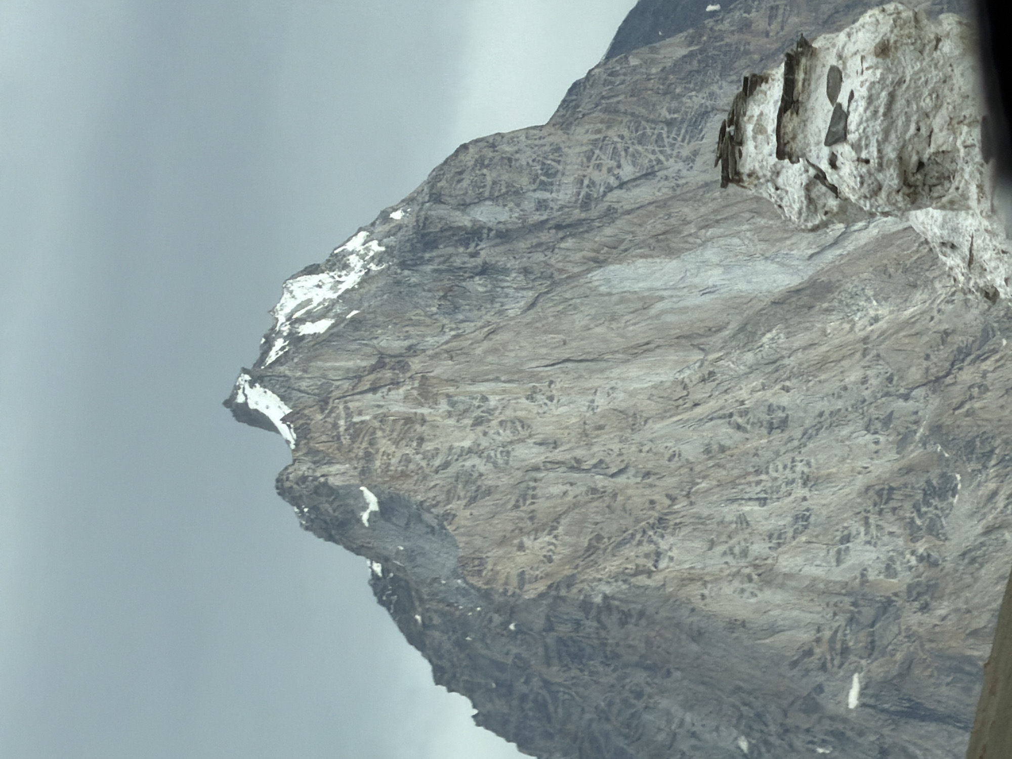 The image shows a close-up of a jagged, grey mountain peak with patches of snow near its summit against a cloudy, overcast sky. In the foreground, a stack of white, weathered rocks, appearing almost like a natural sculpture, is partially visible. The larger mountain's face is textured with vertical striations and appears to be made of rock. There are no people or discernible human activity in the scene. The location is identified as Kurgiakh, India.