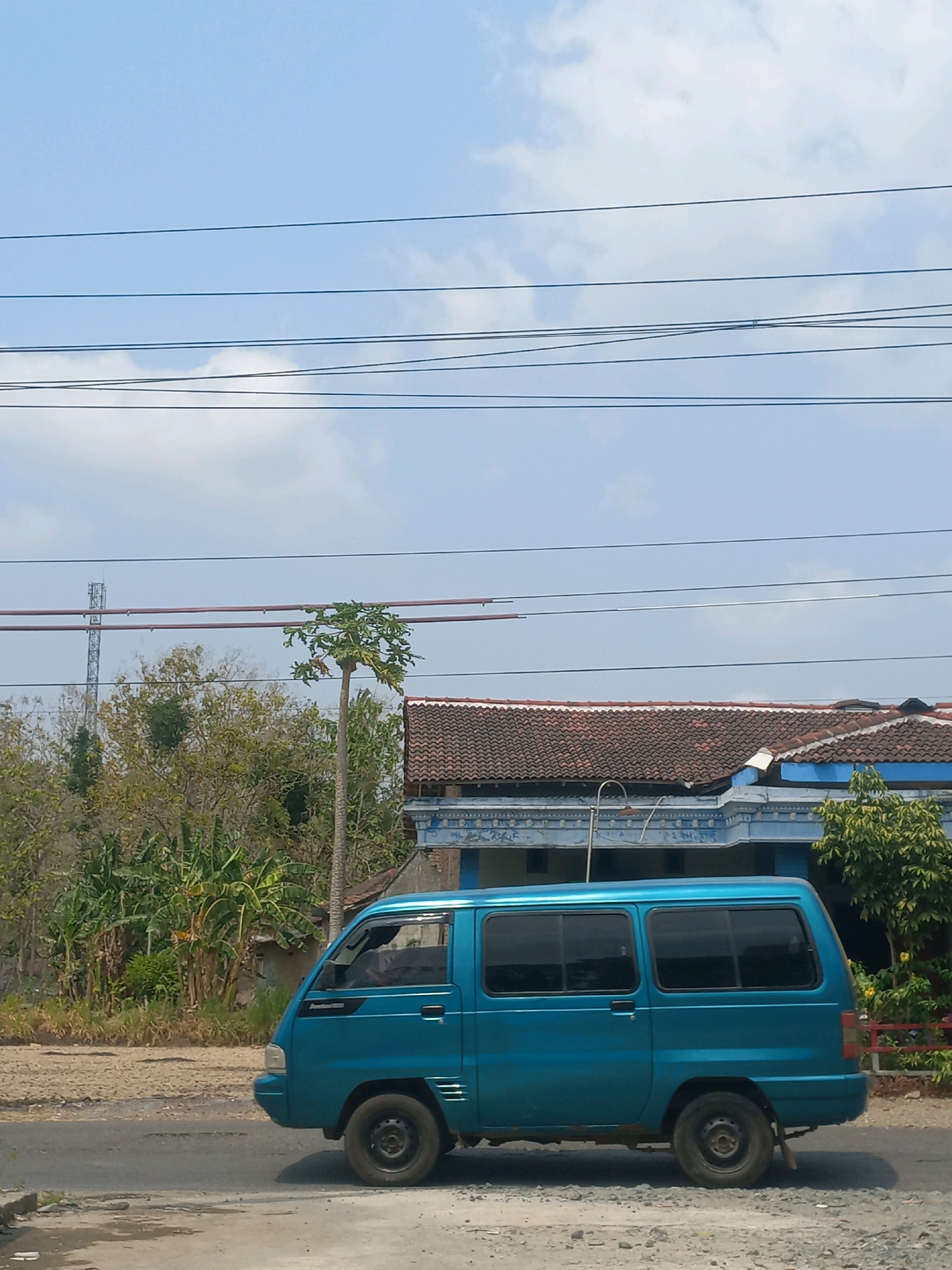 An eye-level, outdoor, medium shot captures a turquoise van parked on a dusty road in Baturetno, Indonesia. The van is positioned in the lower half of the frame, facing left, with its side clearly visible. The road surface is a mix of asphalt and dirt, with some gravel scattered around. Beyond the road, a patch of dry, cracked earth leads to a vibrant green area with banana trees and other foliage. A tall, thin papaya tree stands prominently in the mid-ground, with a few green fruits visible. In the background, a traditional house with a tiled roof and blue trim is partially obscured by vegetation. A cellular tower can be seen in the distance, to the left of the papaya tree. Several power lines stretch horizontally across the upper portion of the image, against a clear blue sky with sparse white clouds. The overall impression is a quiet, rural scene under a bright sky.