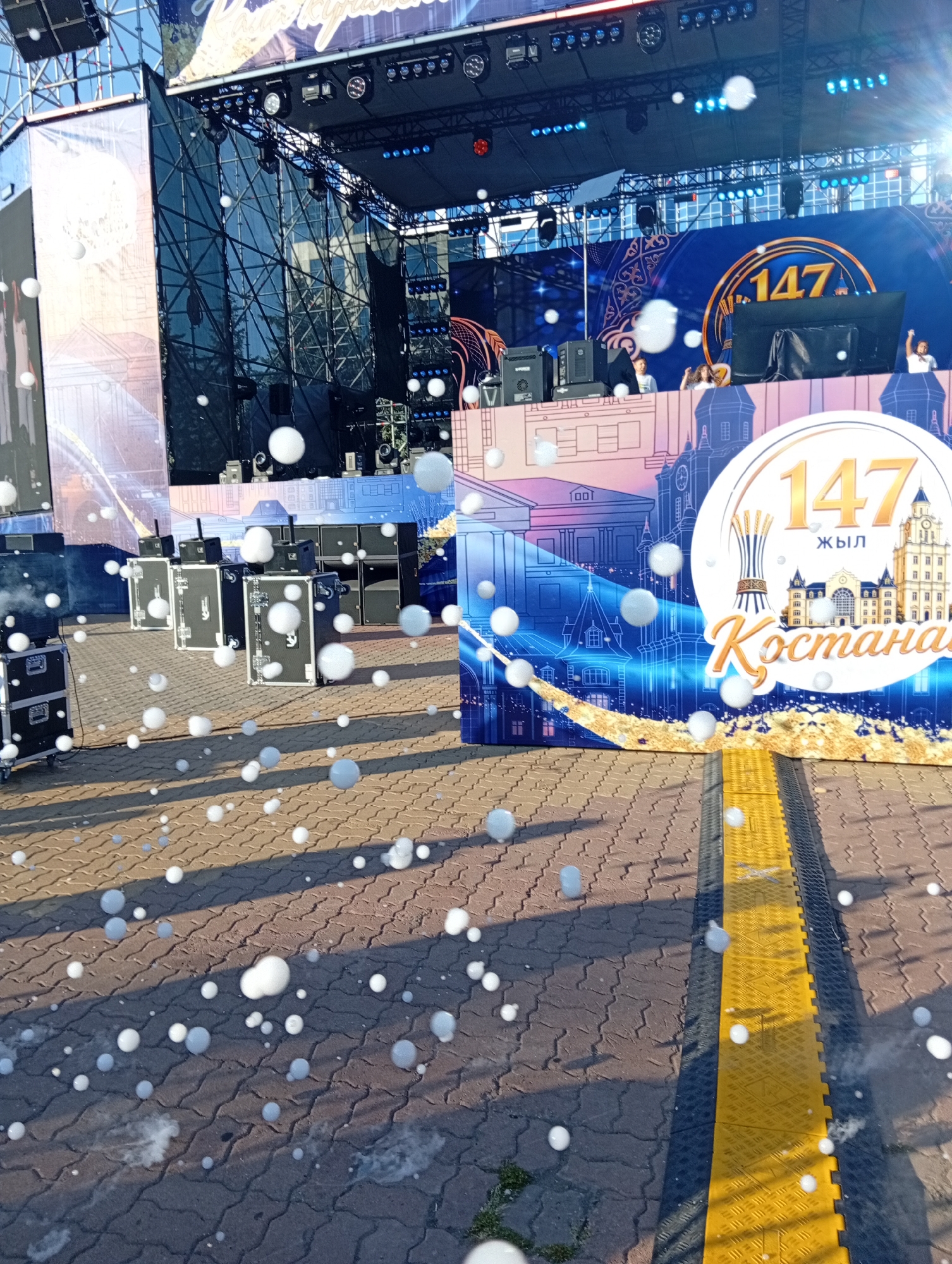 The image depicts an outdoor event stage set up on a paved surface. The stage backdrop features a blue design with golden accents, displaying the text "147 ЖЫЛ ҚОСТАНАЙ" along with images of buildings and what appears to be a harvest symbol. Several large speakers and audio equipment cases are visible on the ground in front of the stage. Many white, spherical objects, resembling soap bubbles, are scattered throughout the foreground and midground of the image, some appearing to be floating in the air and others resting on the ground. The ground itself is paved with interlocking bricks. A yellow and black striped barrier runs across the right side of the image. The lighting suggests it is daytime, with shadows cast on the ground.
