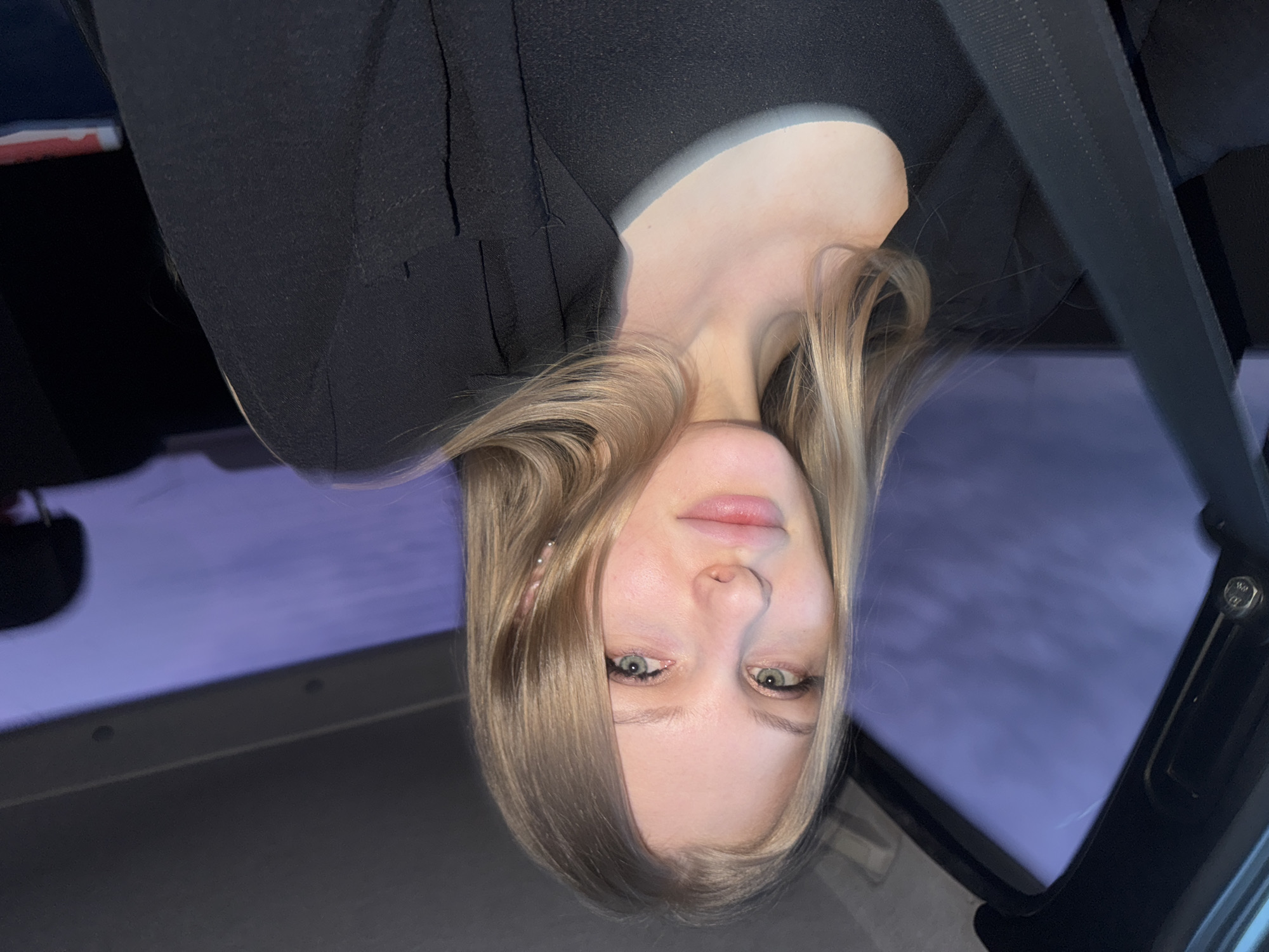The image shows a woman with blonde hair, green eyes, and fair skin. She is wearing a black blazer and a seatbelt. The background appears to be the interior of a vehicle, with a glimpse of a purple-toned outdoor scene visible through the windows. No information suggests that this image was taken in the city of Perm, Russia.