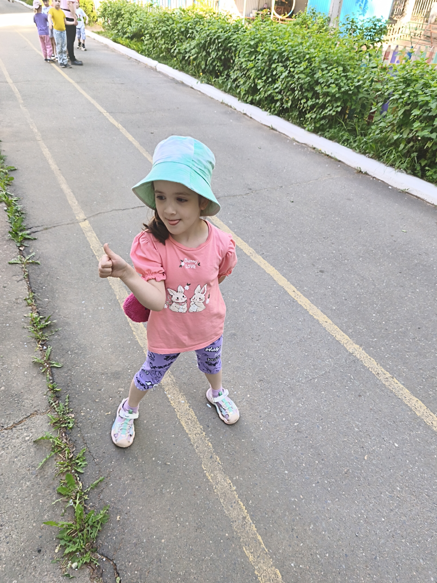 A young girl stands on an asphalt path, giving a thumbs-up. She wears a light blue hat, a pink t-shirt with bunny graphics, purple leggings, and sandals. A pink, fuzzy item is slung over her shoulder. A line of children and adults is visible in the background. The path has yellow painted lines. Green foliage lines the edge of the path. The location is in the city of Chaikovsky, Russia.
