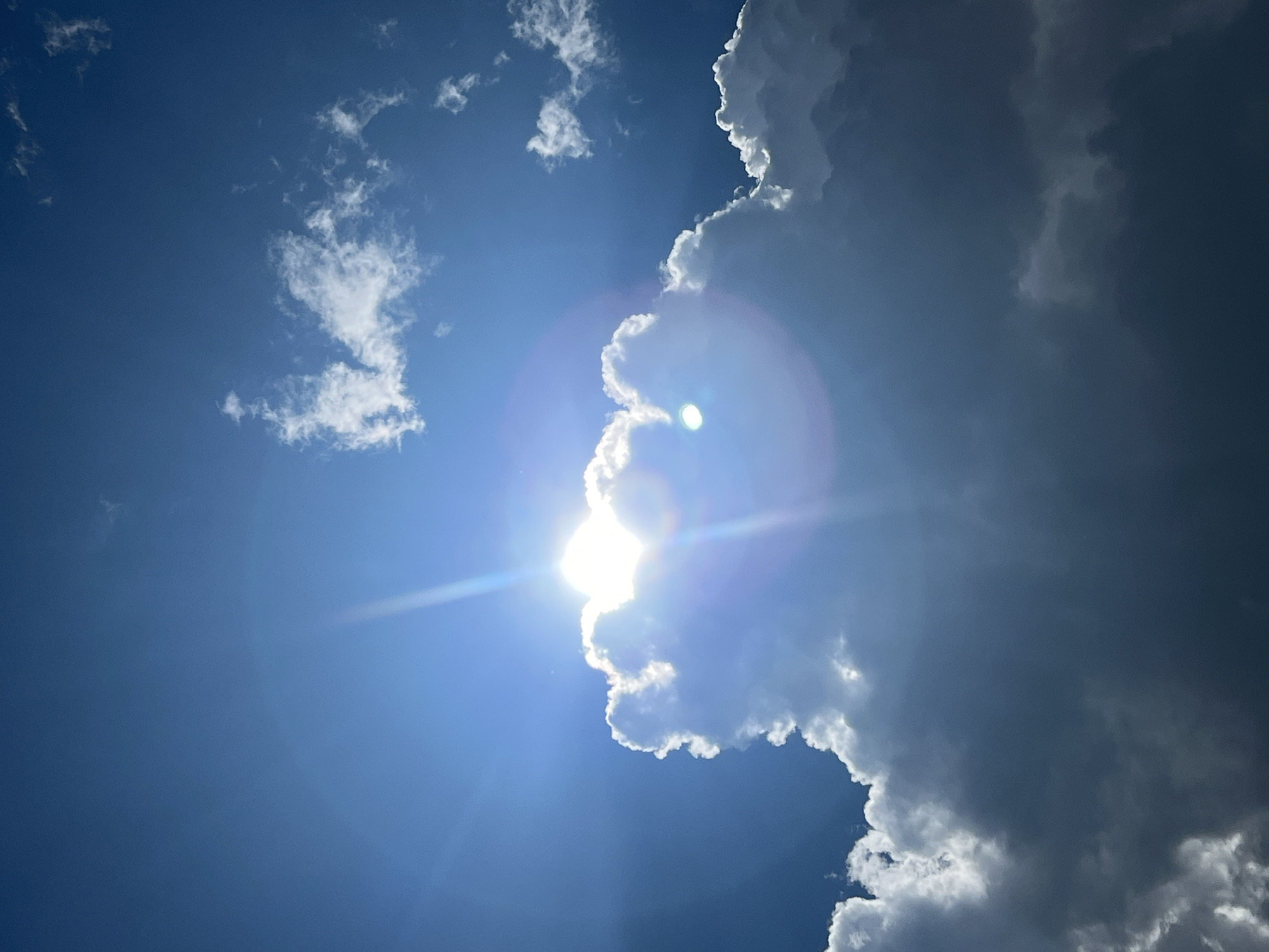 A photograph captures a dramatic sky with a brilliant sun peeking from behind a large, textured cloud. The sun's rays pierce through the cloud, creating a bright glare and lens flare effect. The sky is a deep blue, with smaller, wispy clouds scattered in the upper right portion of the frame. The lower part of the image is dominated by the massive cloud, which is dark and shadowed in places, indicating its thickness and depth. The photograph appears to be taken from a ground-level perspective, looking upwards. The scene is set in Mochishche, Russia, on a day with dynamic cloud formations and strong sunlight. There are no people or man-made objects visible in the image.