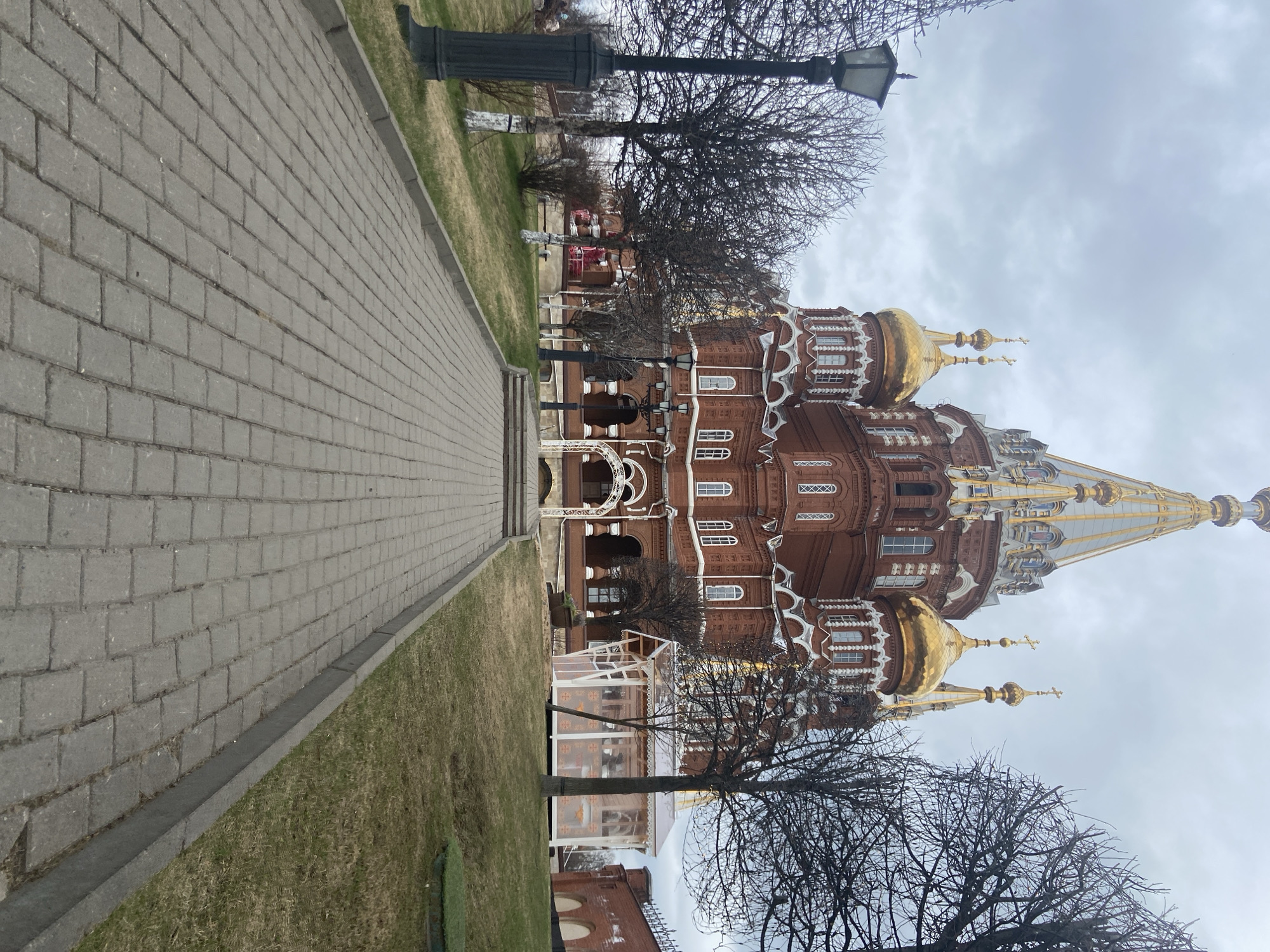 A brown brick building with golden domes is located on a street with gray brick sidewalks. A black lamppost and bare trees are present. The sky is overcast. The location is in the city of Unknown, Unknown.