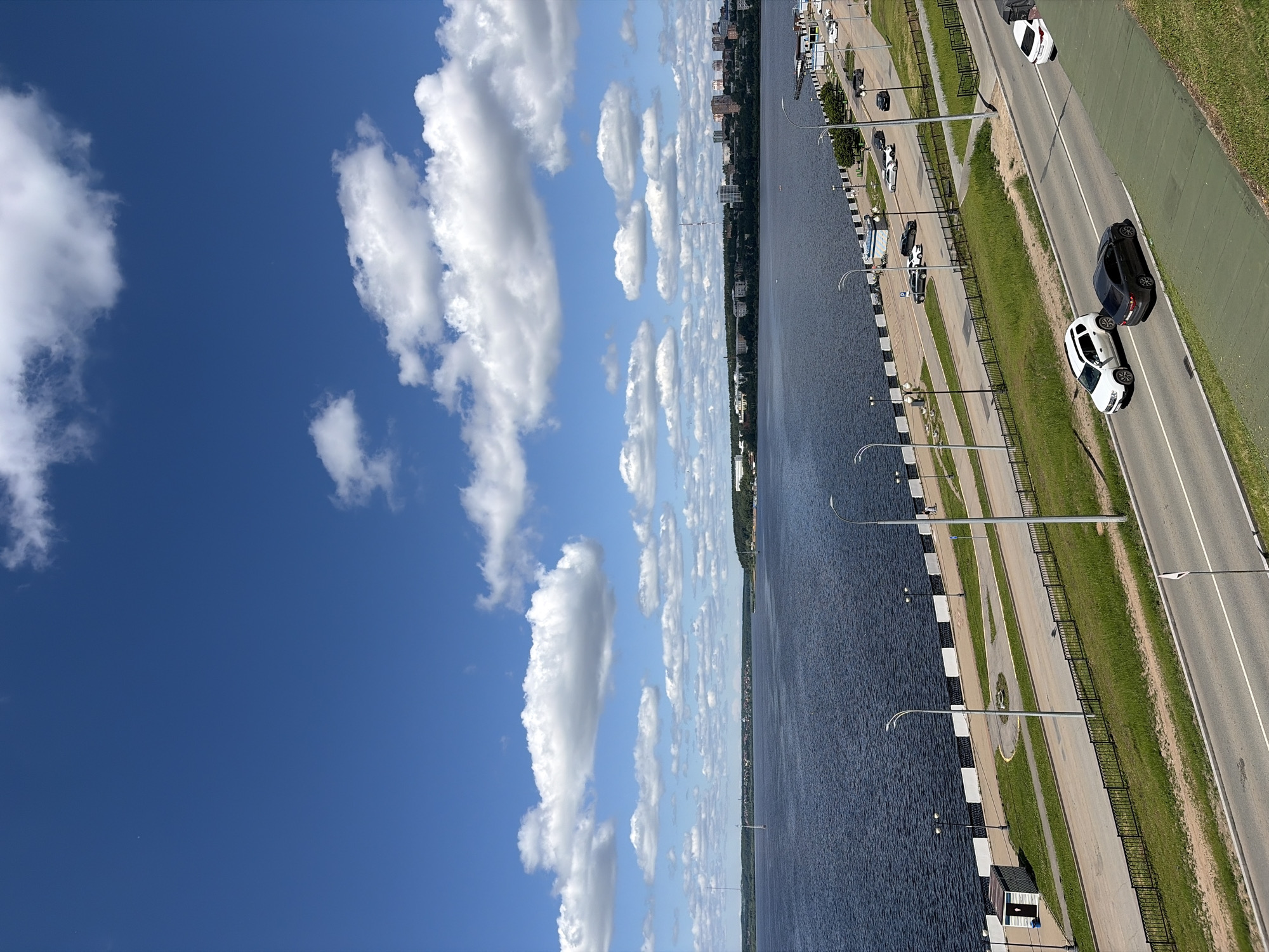 The image, when correctly oriented, presents a high-angle panoramic view of a wide body of water, likely a river or reservoir, under a bright blue sky with scattered white cumulus clouds. This scene depicts a waterfront area in Izhevsk, Russia.
The upper portion of the frame is dominated by the clear, deep blue sky adorned with well-defined, fluffy white clouds. The body of water, a dark blue, extends across the middle of the image, appearing calm with subtle ripples reflecting the daylight.
On the right side of the image, running parallel to the water, is a developed embankment. It features a paved walkway equipped with modern, curved lampposts and a black metal fence separating the walkway from the water. Inland from the walkway, a strip of green grass leads to a two-lane asphalt road with clear white lane markings. Several vehicles are visible on the road; a white SUV and a black SUV are prominent in the foreground, seemingly in motion away from the viewer. Other cars are visible further down the road, with some potentially parked along the embankment.
The distant left bank of the water body reveals a continuous urban landscape, featuring various buildings and structures interspersed with trees and greenery, forming a city skyline along the horizon. The scene takes place on a clear, sunny day, likely during midday or early afternoon, given the bright illumination. No specific human figures are distinctly visible, and no discernible text is present in the image. The overall impression is one of a serene yet active urban waterfront.