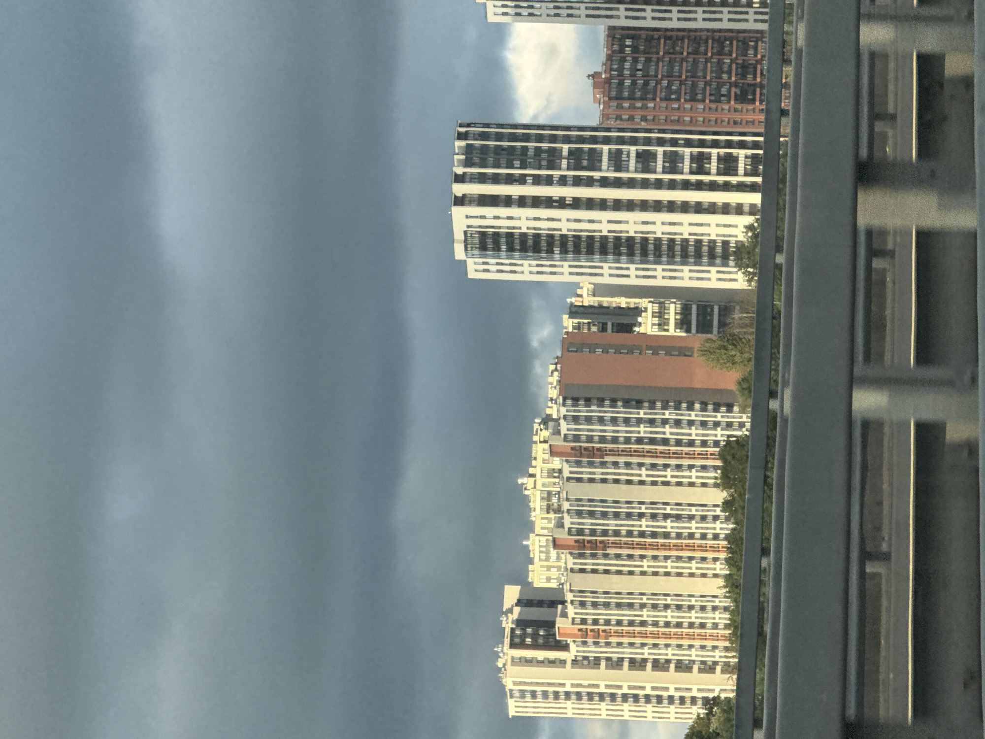 A wide shot captures a line of modern, high-rise residential buildings against a dark, cloudy sky. The buildings are a mix of beige, white, and reddish-brown facades, with numerous windows creating a repetitive pattern. The architecture suggests a contemporary urban development. In the foreground, a gray highway barrier with horizontal railings runs across the bottom of the frame, obscuring the lower portion of the buildings and the ground. The scene appears to be taken from a moving vehicle on the highway, looking towards the city. The lighting is diffused due to the overcast sky, casting a somewhat muted atmosphere over the scene. This appears to be the area of the city of Saint Petersburg, Russia, possibly near the Gulf of Finland, given the characteristic architecture and the presence of large residential complexes. There are no people visible in the image.