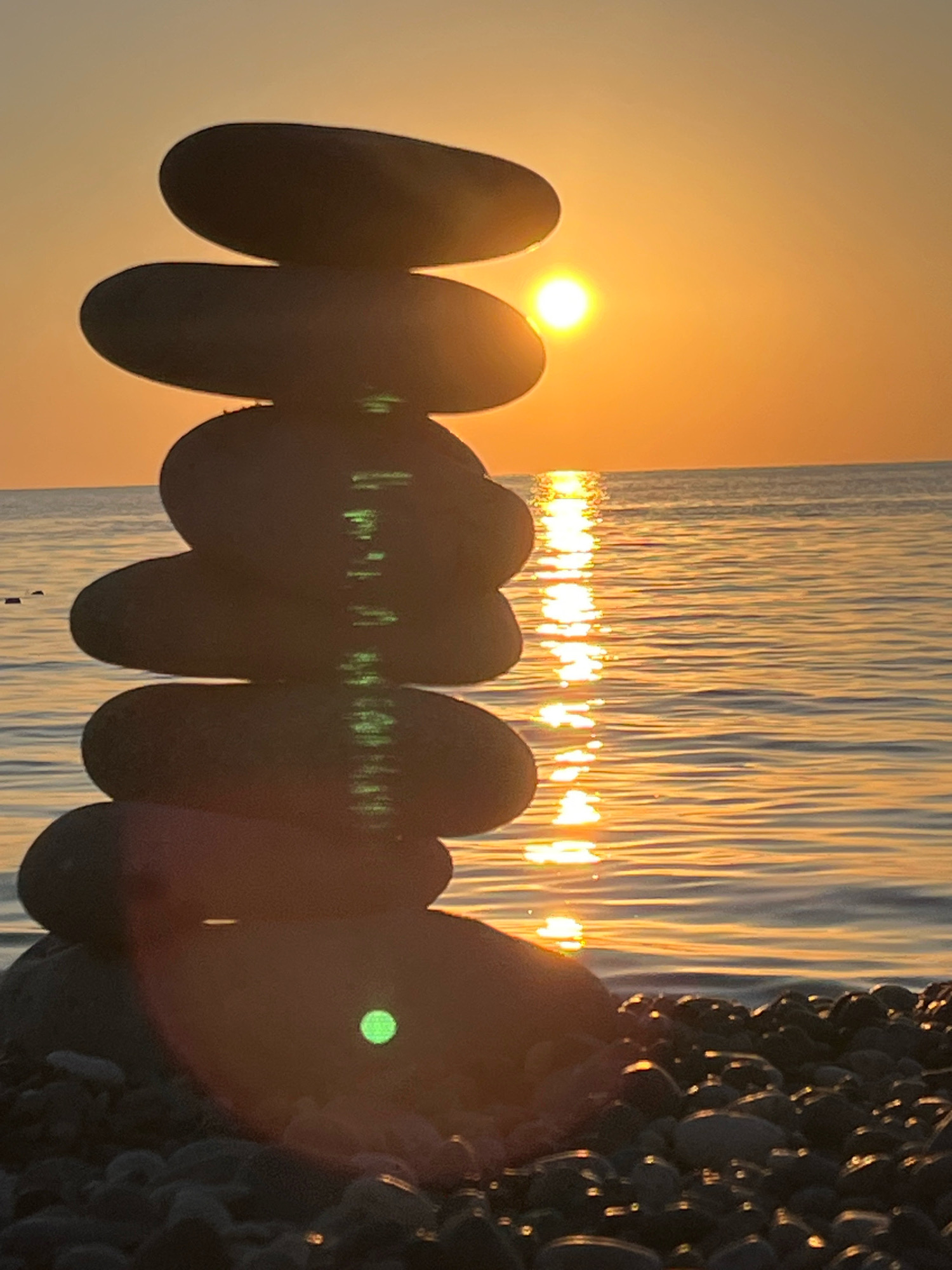 A stack of five dark, smooth stones is balanced on a pebble beach. The stones are arranged vertically, with the largest at the bottom and progressively smaller stones stacked on top. The sun is setting over a calm body of water, casting a golden reflection on the surface. The sky is a gradient of orange and yellow. The scene is located on the coast of Kobuleti, Georgia.
