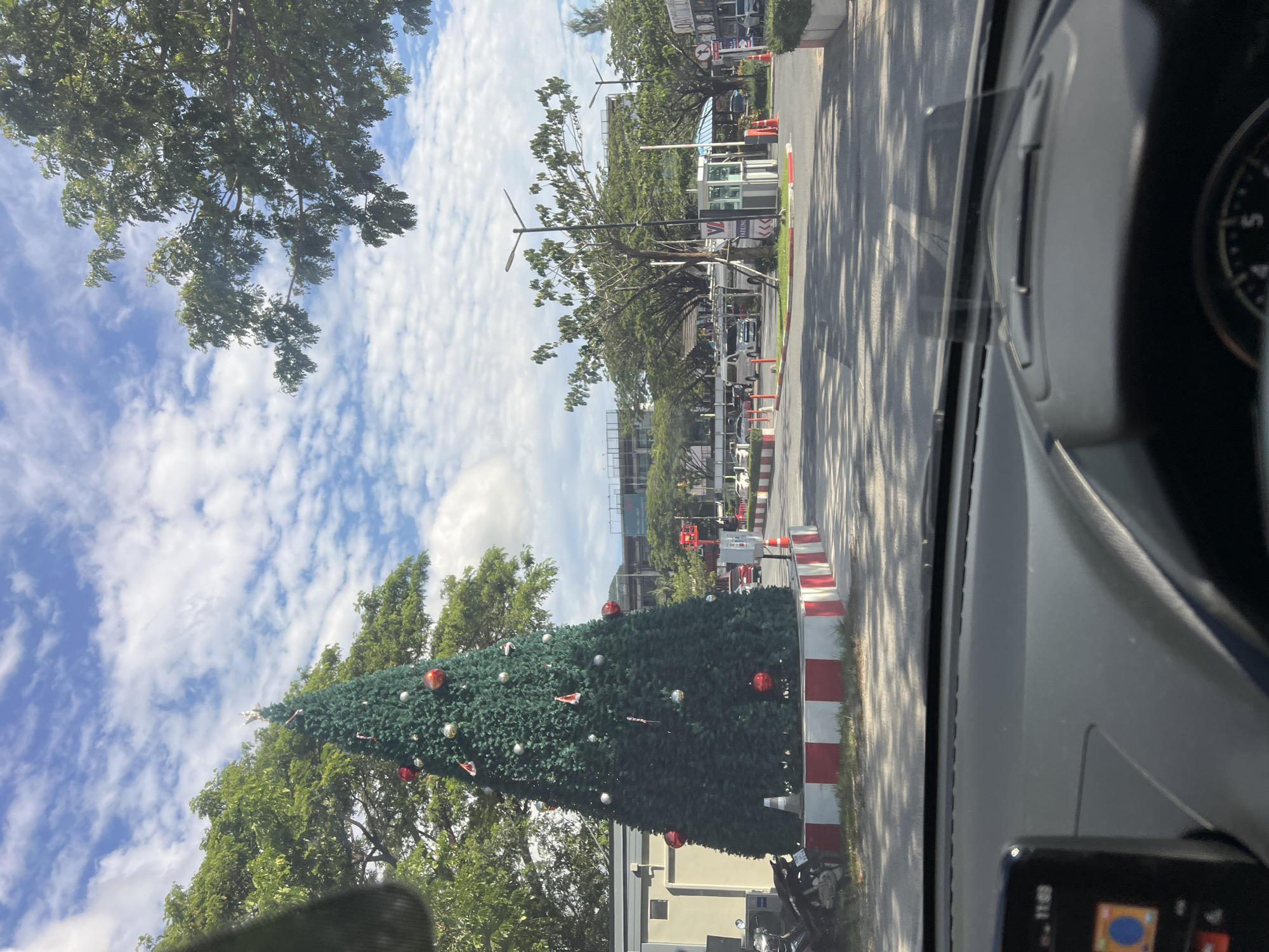 The image captures a daytime outdoor scene, likely in a city environment, with a large, decorated Christmas tree prominently displayed. The perspective is from inside a vehicle, looking out through the windshield and dashboard.
The Christmas tree is a tall, conical evergreen, adorned with red and silver ornaments. It stands on a raised circular platform with red and white striped borders. Surrounding the tree are green trees and foliage. In the background, there are buildings, streetlights, traffic cones, and what appears to be a parking area with several vehicles including a white van, cars, and motorcycles. A guard booth or information kiosk is visible near the entrance to the parking area. The sky is blue with scattered white clouds. The road surface is asphalt, with painted white lines indicating lanes. The dashboard of the vehicle, including a portion of the steering wheel and a screen displaying time and what looks like GPS information, is visible in the foreground.