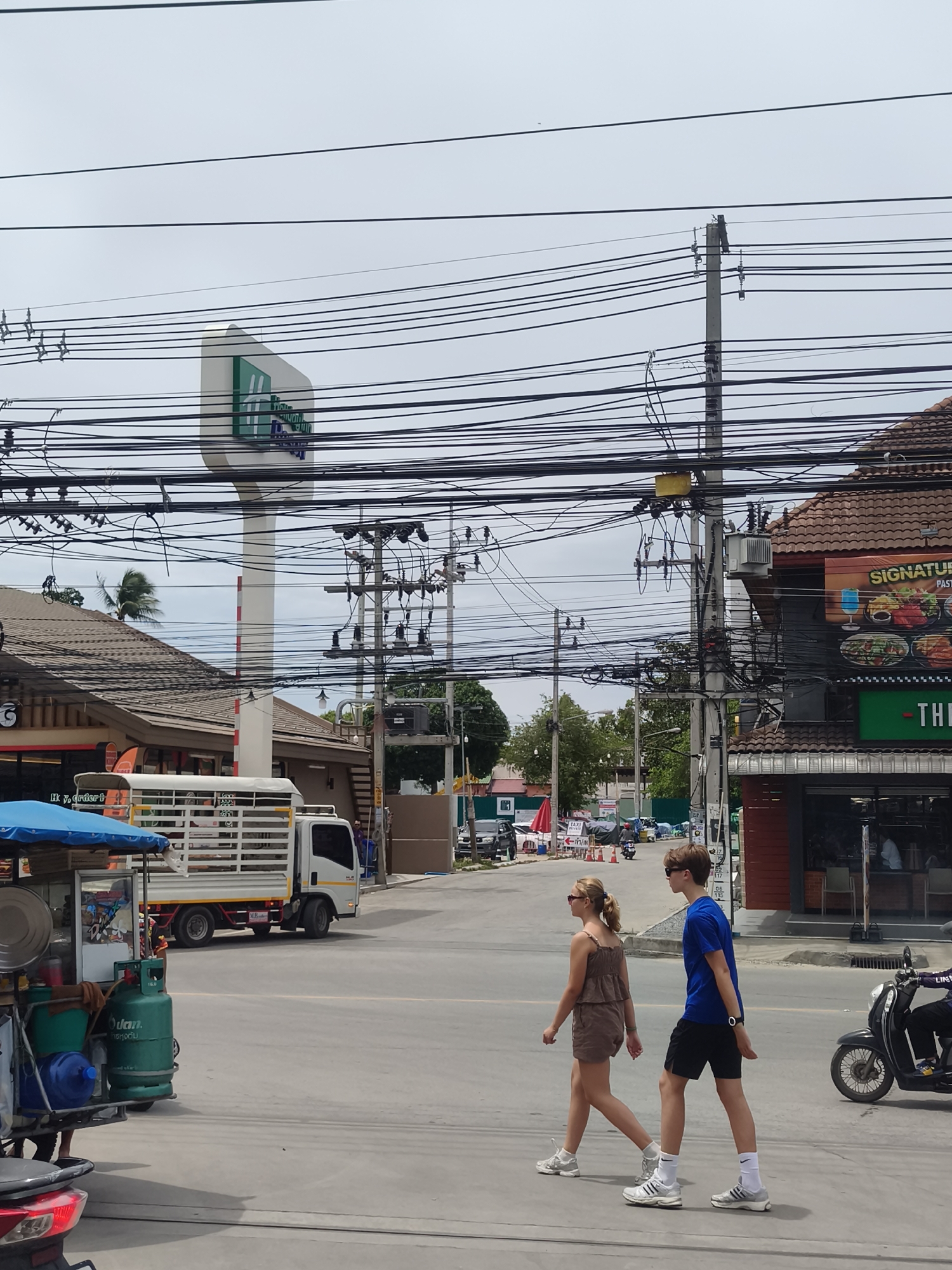 Two young individuals, a female and a male, are walking across a paved road in an urban environment, likely Ko Samui District, Thailand. The female, with blonde hair pulled back, is wearing a brown sleeveless romper and white athletic shoes. The male, with light brown hair and sunglasses, is dressed in a blue t-shirt, black shorts, white socks, and white athletic shoes. They are walking side-by-side, appearing to be engaged in conversation or simply walking together.
The background reveals a street scene with various elements. Overhead, a dense network of black electrical wires crisscrosses the sky, supported by utility poles. To the left, a tall white sign with a green logo, possibly for Holiday Inn, stands prominently. Adjacent to it is a building with a brown tiled roof and brown siding. A white truck with a cage-like bed is parked on the street. In the foreground, a mobile food cart with a blue awning is visible, equipped with cooking utensils, a gas cylinder, and water containers. To the right, another building with a brown tiled roof features a sign that reads "SIGNATURE". A black motorcycle is parked and a person is riding another motorcycle down the street. The sky is overcast, suggesting a possibly humid or cloudy day typical of tropical locations. The road appears to be a main thoroughfare with traffic cones visible further down.