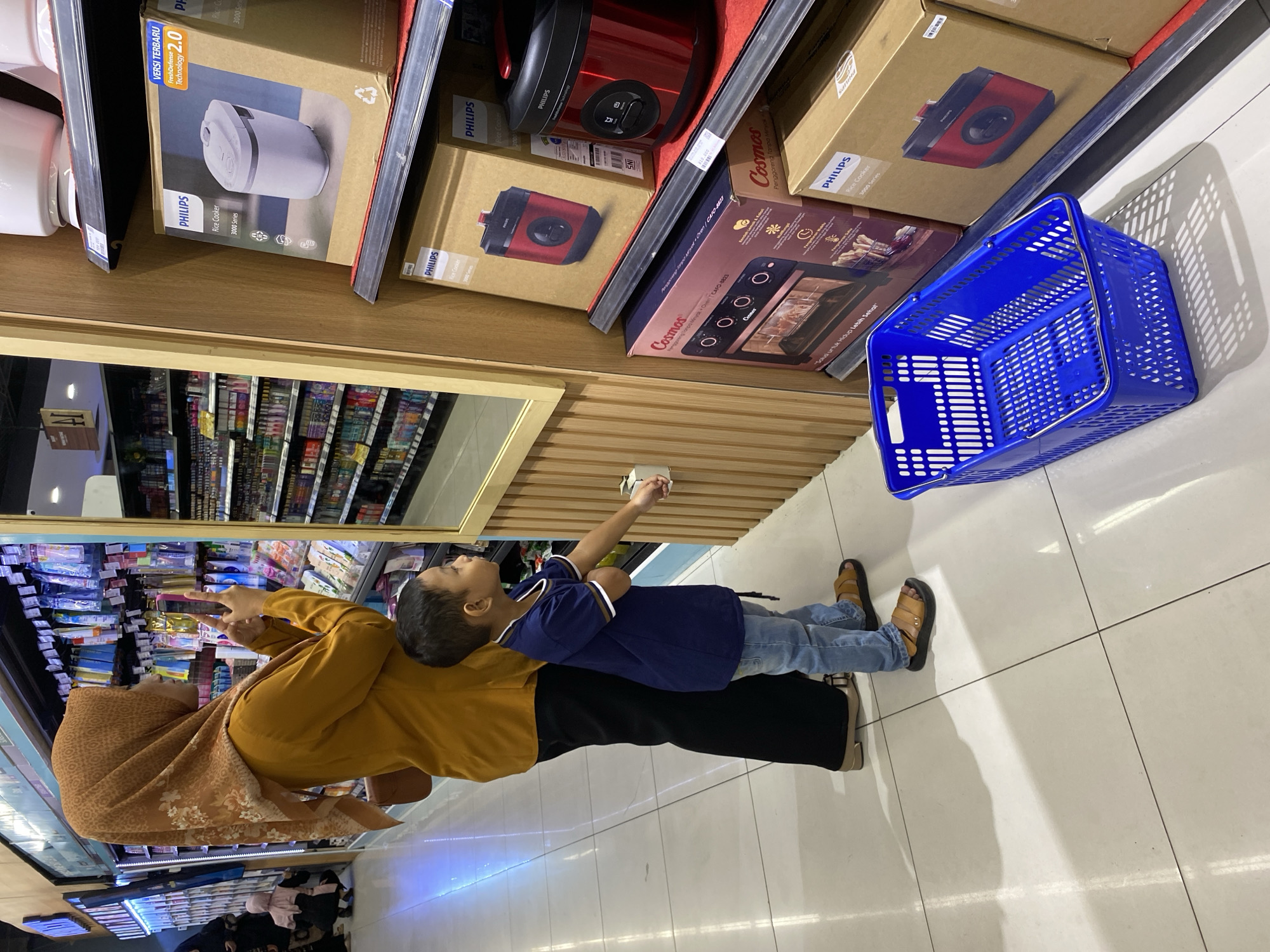 The image depicts a mother and son in a retail environment, likely a supermarket or department store, with shelves stocked with various household appliances. The mother, wearing a yellow tunic and a patterned hijab, is standing in profile, holding a smartphone and appearing to take a photo. Her son, a young boy in a navy blue t-shirt and blue jeans, is standing next to her, reaching out towards a small white object on a shelf.
The shelves are filled with branded products, notably several boxes of Philips rice cookers and a Cosmos brand oven. The environment is well-lit, with a polished floor reflecting the overhead lighting. A blue shopping basket is placed on the floor in the foreground. The presence of signage and the general layout suggest a commercial setting within a city. While the specific location is not explicitly stated in the image, such retail establishments are common in urban centers like Banda Aceh, Indonesia, a city known for its shopping malls and markets. The scene captures a mundane moment of everyday life, possibly during a shopping trip for household goods.