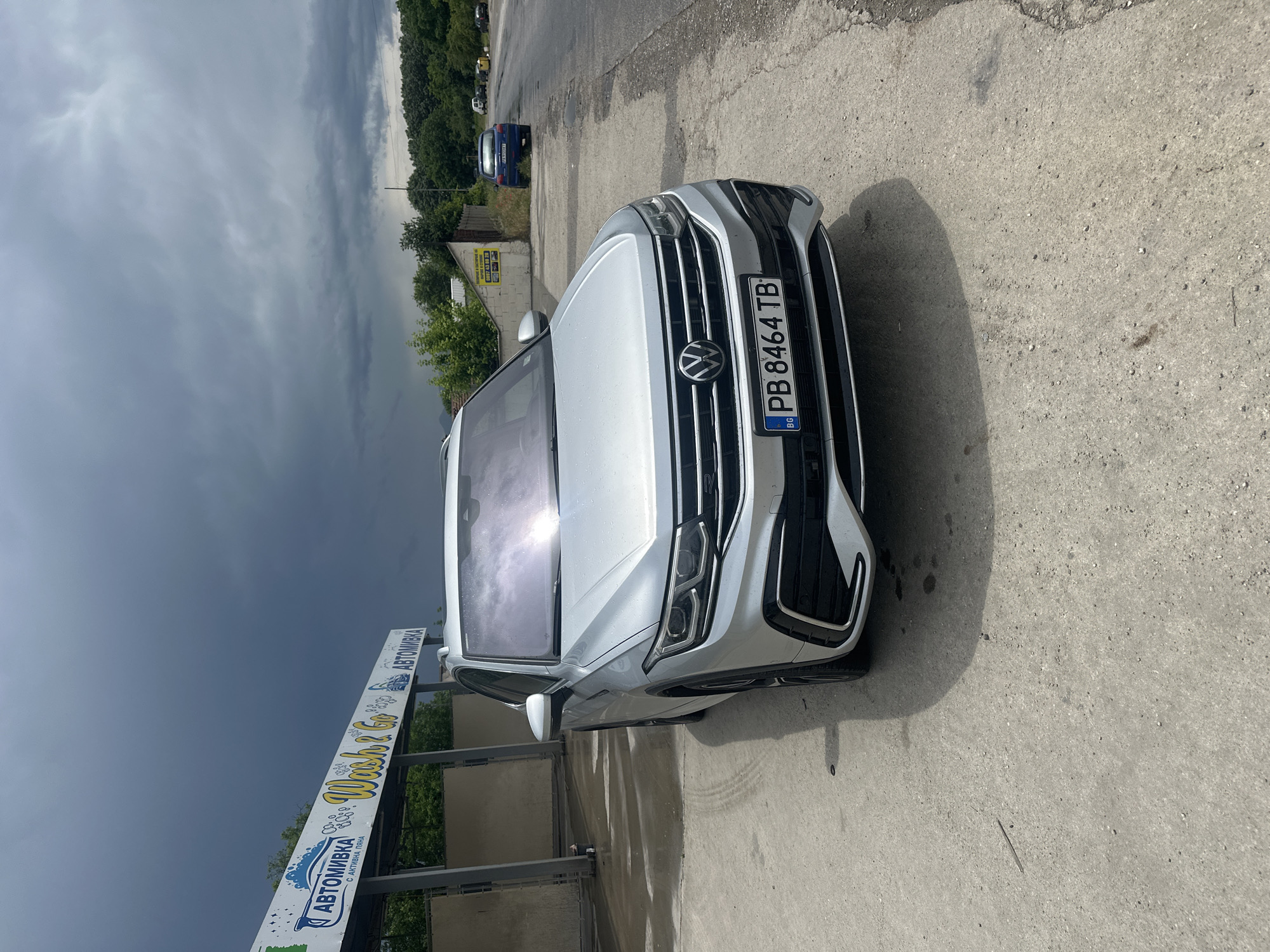 A silver Volkswagen SUV is prominently featured at an outdoor car wash facility. The car, possibly a Tiguan or a similar model, is parked on a concrete surface, appearing wet as if recently washed, with sunlight reflecting off its windshield. Its Bulgarian license plate, clearly visible on the front, reads "PB 8464 TB".
To the bottom left, a section of a car wash sign is visible, displaying text in Cyrillic and English. The Cyrillic text includes "АВТОМИВКА" (Avtomivka), meaning "Car Wash," and "С АКТИВНА ПЯНА" (S Aktivna Pyana), which translates to "With Active Foam." The English part of the sign shows "Wash & Go" in a decorative script.
The background is dominated by a dramatic, overcast sky with dark clouds, hinting at recent rain or an impending storm. Beyond the car wash structure and sign, there's a line of green trees. Further into the distance, a blue car is parked on what appears to be a dirt or gravel path, with more trees and a small building visible behind it. The ground in the foreground is concrete, showing some puddles and wet marks consistent with a car wash environment. The setting is Ivailo, Bulgaria, indicated by the license plate and the language on the sign. The image captures a daytime scene.