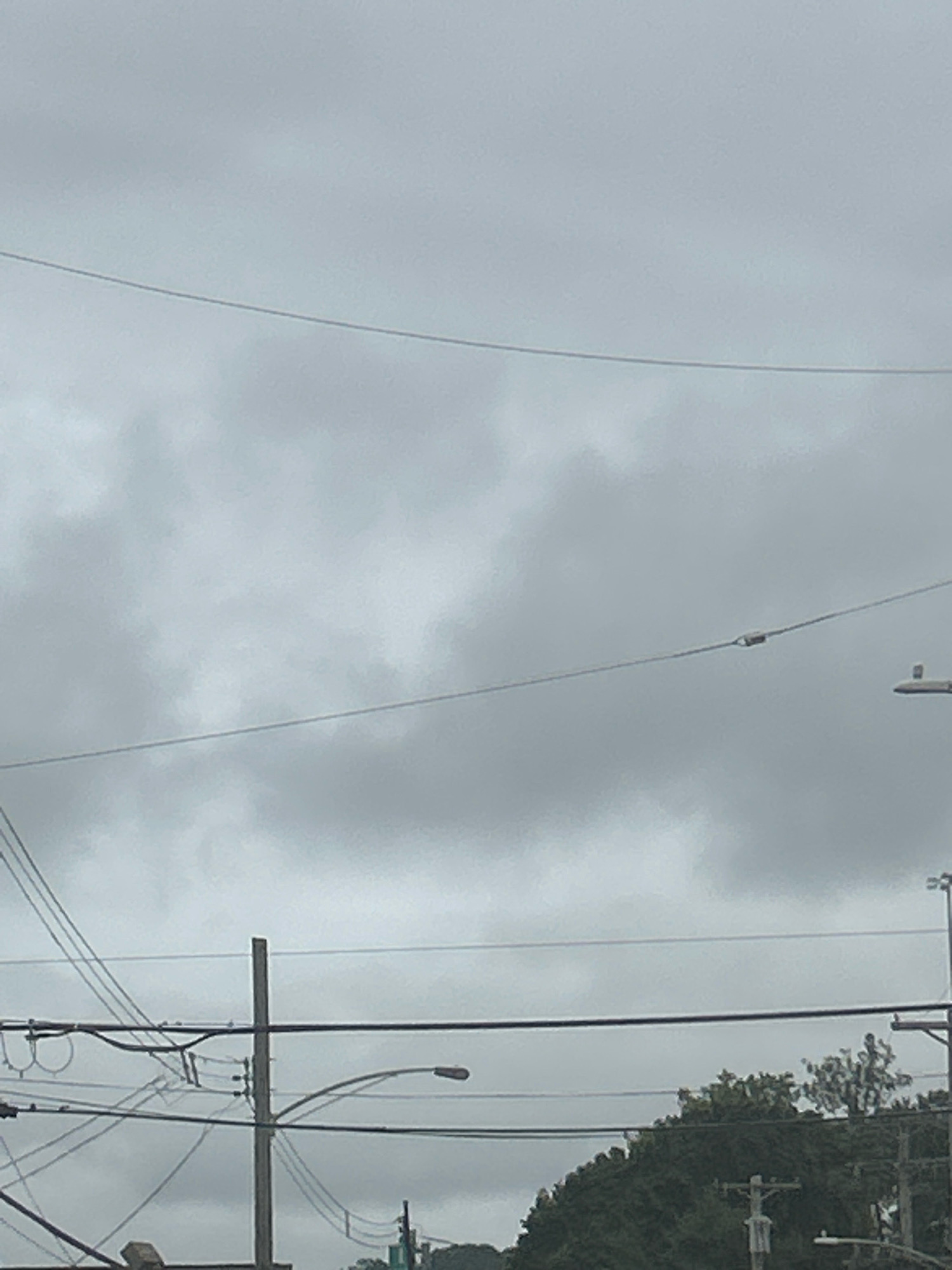 The image captures an overcast sky with visible clouds, crisscrossed by utility wires. Several utility poles are present, supporting the network of wires. In the lower portion of the image, there is a streetlight and a dense cluster of trees. The scene appears to be an outdoor urban or suburban environment.