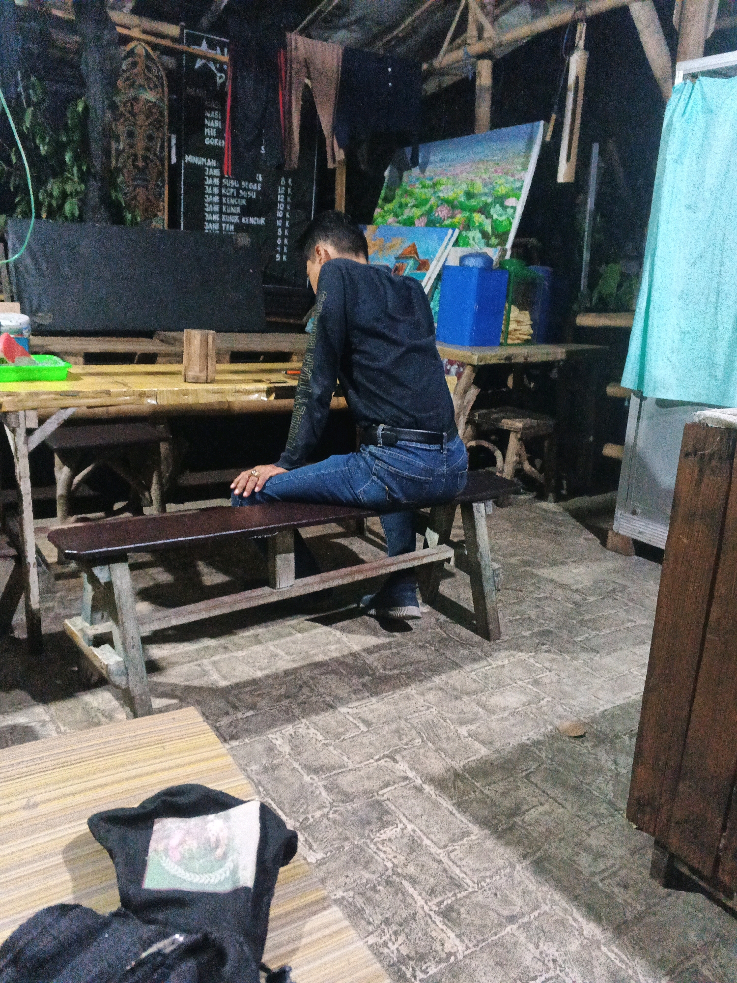 The image depicts an outdoor seating area, possibly a warung or street-side cafe, at night in Ungaran, Indonesia. The scene is dimly lit, with bamboo structures forming part of the roof and walls. A man, wearing a dark long-sleeved shirt and blue jeans, is seated on a wooden bench. He is facing away from the camera, with his legs crossed at the knee and his hands resting on his lap. A ring is visible on his right hand.
In the background, a menu board is partially visible, listing food and drink items such as "Nasi Goreng" and various "Jamu" (traditional Indonesian herbal drinks). Several paintings are propped up, one of which depicts a landscape with water and flowers. Blue plastic containers and other items are scattered around the tables, which are made of bamboo and wood. The ground is paved with bricks.
In the foreground, a black hooded jacket with a printed image on the back is lying on a wooden surface, likely a table. The overall ambiance suggests a casual, rustic setting.