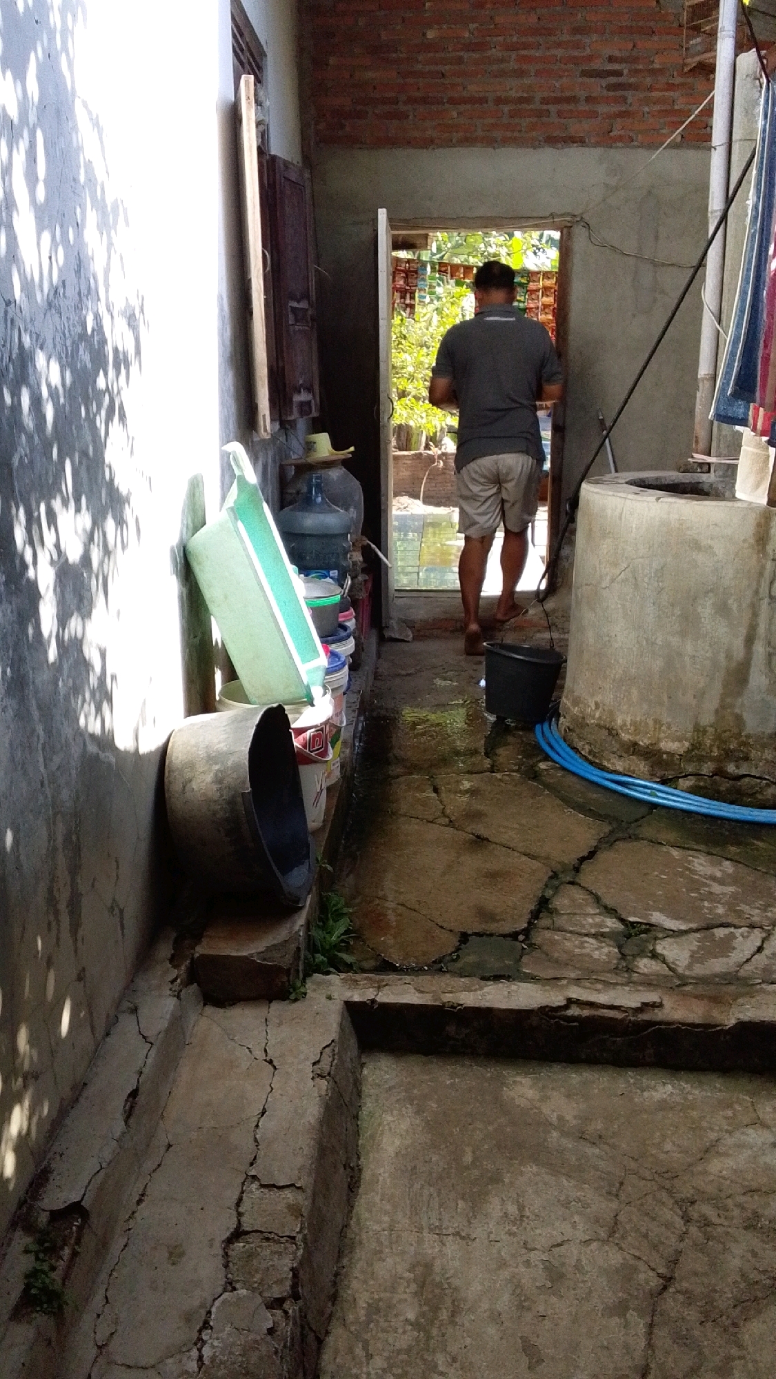 A man wearing a grey t-shirt and light-colored shorts walks through an open doorway into a sunlit area. He is facing away from the camera. The scene appears to be an outdoor alleyway or courtyard. To the left, a white wall is illuminated by dappled sunlight. Stacked on a ledge against the wall are several large water jugs and plastic basins, including a prominent green basin. To the right of the doorway is a large, cylindrical concrete structure, possibly a well, with blue tubing coiled around its base. The ground is paved with cracked concrete, and there are puddles of water on the surface. In the background, lush green foliage is visible through the doorway. The architecture suggests a residential or utilitarian setting in Lawu, Indonesia.