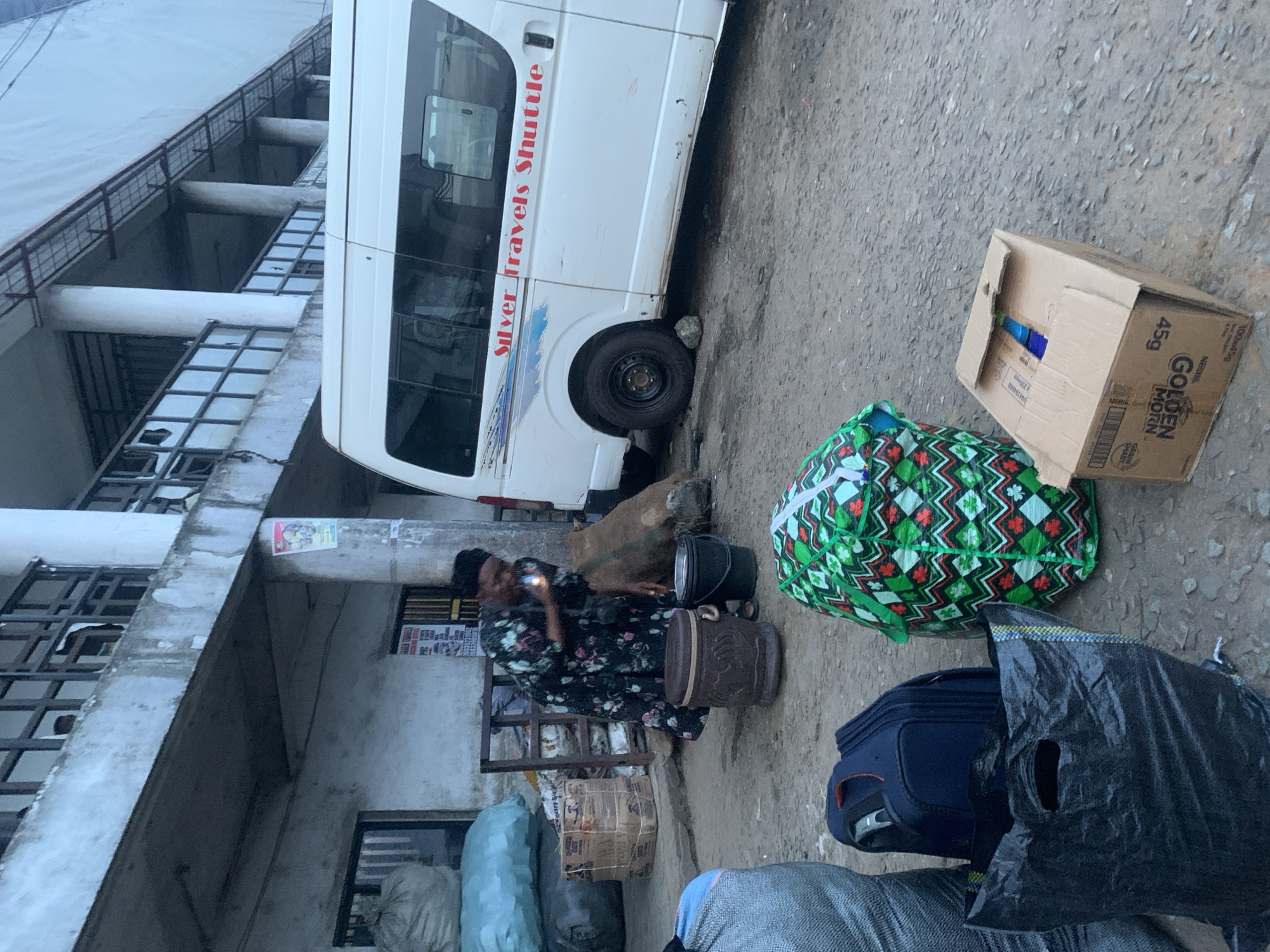 In the city of Elelenwo, Nigeria, a scene unfolds outside a building. A woman, dressed in a dark floral patterned dress and a headscarf, is bent over, holding a small object that appears to be a lighter near her face. She is standing on a dirt and gravel surface. In front of her are several objects: a dark brown, patterned bucket with a handle, and a black bucket. To the right of the buckets, a large sack made of rough material is partially visible. Behind the woman, a white shuttle bus with red lettering that reads "Silver Travels Shuttle" is parked. The bus has tinted windows and a silver stripe running along its side. Various bags and luggage are scattered around the scene. To the left of the woman, large, light blue and grey sacks are stacked. In the foreground, a navy blue suitcase and a large, green and white patterned bag are placed on the ground. A black plastic bag with handles is also visible in the foreground. Behind the shuttle bus, a large, rough sack is propped against it. A cardboard box with visible labels, including "GOLDEN MORN 45g", is also on the ground in the foreground. The environment appears to be an open area, possibly a bus station or market, with a building characterized by concrete columns and balconies in the background. The lighting suggests it is daytime, possibly overcast.
