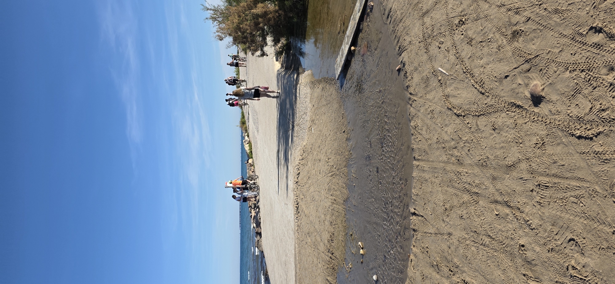 The image shows a coastal scene in Canet d'en Berenguer, Spain. A concrete path runs along the edge of the beach, separating the sand from a shallow stream of water flowing into the sea. The sky is clear blue. Several people are walking and cycling along the path. A few others are near the water’s edge. The sand is marked with tire tracks and footprints. A small bush or tree is present near the stream.