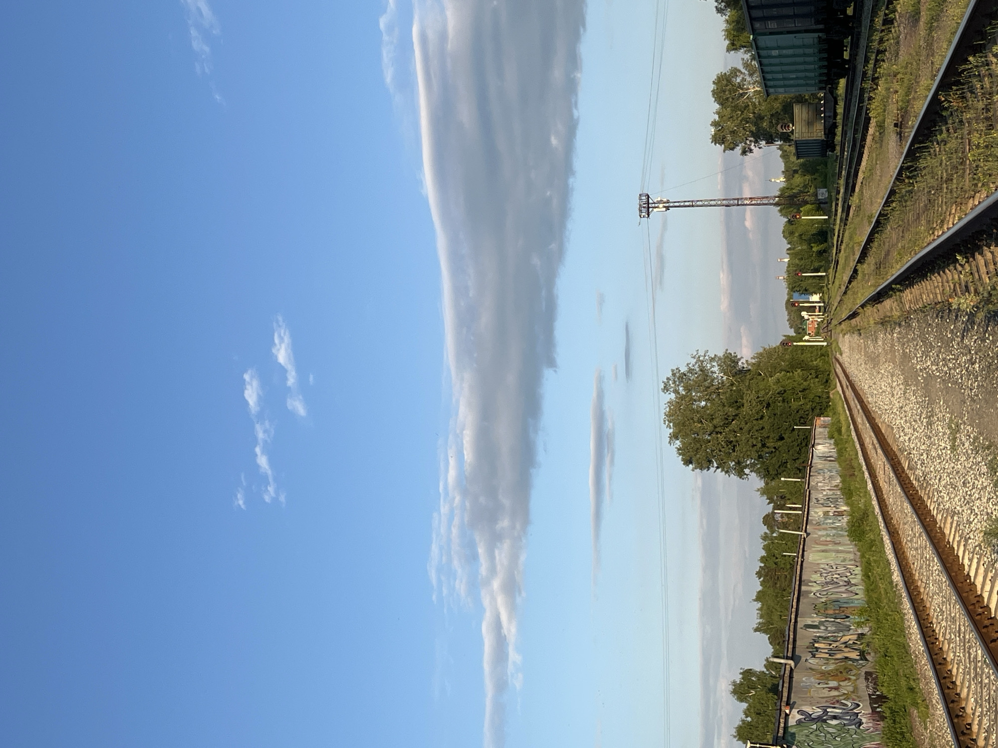 This is a wide-angle, eye-level shot of a railway yard under a clear blue sky with scattered white clouds. In the foreground, two sets of railway tracks run parallel towards the horizon. The tracks are made of metal rails and gravel. To the left of the tracks, a long concrete wall is covered in graffiti. Beyond the wall, a dense line of trees forms a green backdrop. To the right of the tracks, several train cars are visible, including a green hopper car. A tall, slender metal tower with electrical equipment stands in the middle ground, and a few distant buildings with white spires can be seen amongst the trees on the horizon. The lighting suggests it is late afternoon or early evening, with soft sunlight illuminating the scene. The location is the Chaikovsky railway station in Chaikovsky, Russia. There are no people or animals visible in the image.