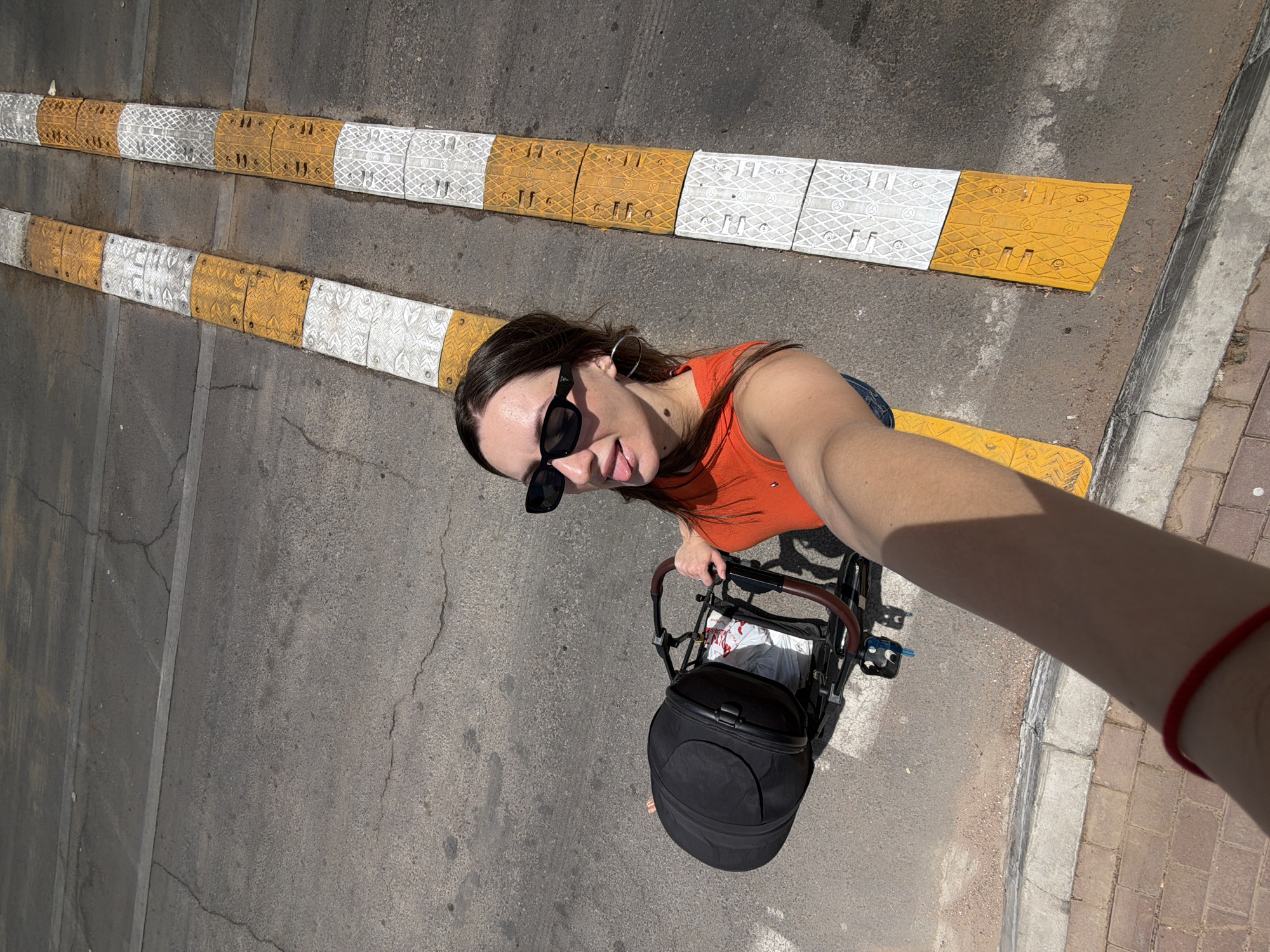 A woman is taking a selfie outdoors, in what appears to be a city environment. She is wearing a bright orange sleeveless top and black sunglasses, with her tongue sticking out playfully. She has on hoop earrings and a red string bracelet on her left wrist. In her right hand, she holds a stroller, which has a large black bag attached to it. The stroller appears to be a lightweight model.
The background shows a cracked asphalt road surface. Two parallel speed bumps, made of alternating yellow and white reflective panels, run along the right side of the image. The speed bumps are textured and appear to be recently installed or maintained. The overall lighting suggests it is daytime, with the sun casting shadows.
Based on the visual cues, such as the style of the speed bumps and the general urban environment, this location is consistent with Sharm El Sheikh, Egypt. Specifically, the reflective speed bumps with their patterned panels are common in tourist areas and along roads in Sharm El Sheikh. The dry, sunny climate is also characteristic of the region.