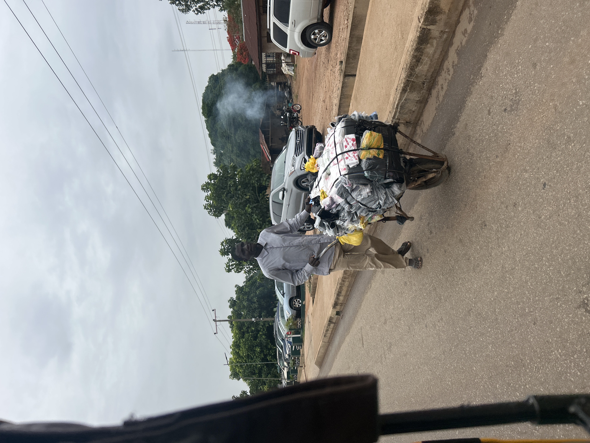 A man is pulling a heavily laden hand cart along a road in Jos, Nigeria. The cart is filled with packaged goods, possibly for sale. The man is wearing a light-colored long-sleeved shirt and tan pants. The road is paved and has a curb on the right. Cars are parked along the road. In the background, there is a small cluster of buildings and green vegetation. Smoke rises from an unknown source in the background. Overhead power lines are visible in the sky.