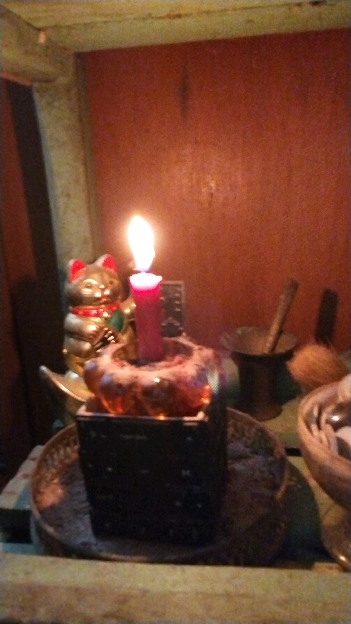 A dimly lit still life setup is depicted, featuring a lit red candle in a decorative holder. Behind the candle, a golden Maneki-neko figurine with red ears and a red collar is visible. To the right of the candle, a small mortar and pestle, and a coconut are present. The scene appears to be situated indoors, possibly on a shelf or altar, with a textured reddish-brown background. The overall impression is one of a spiritual or ritualistic setting. No specific location in Penompo, Indonesia can be determined from the image alone.