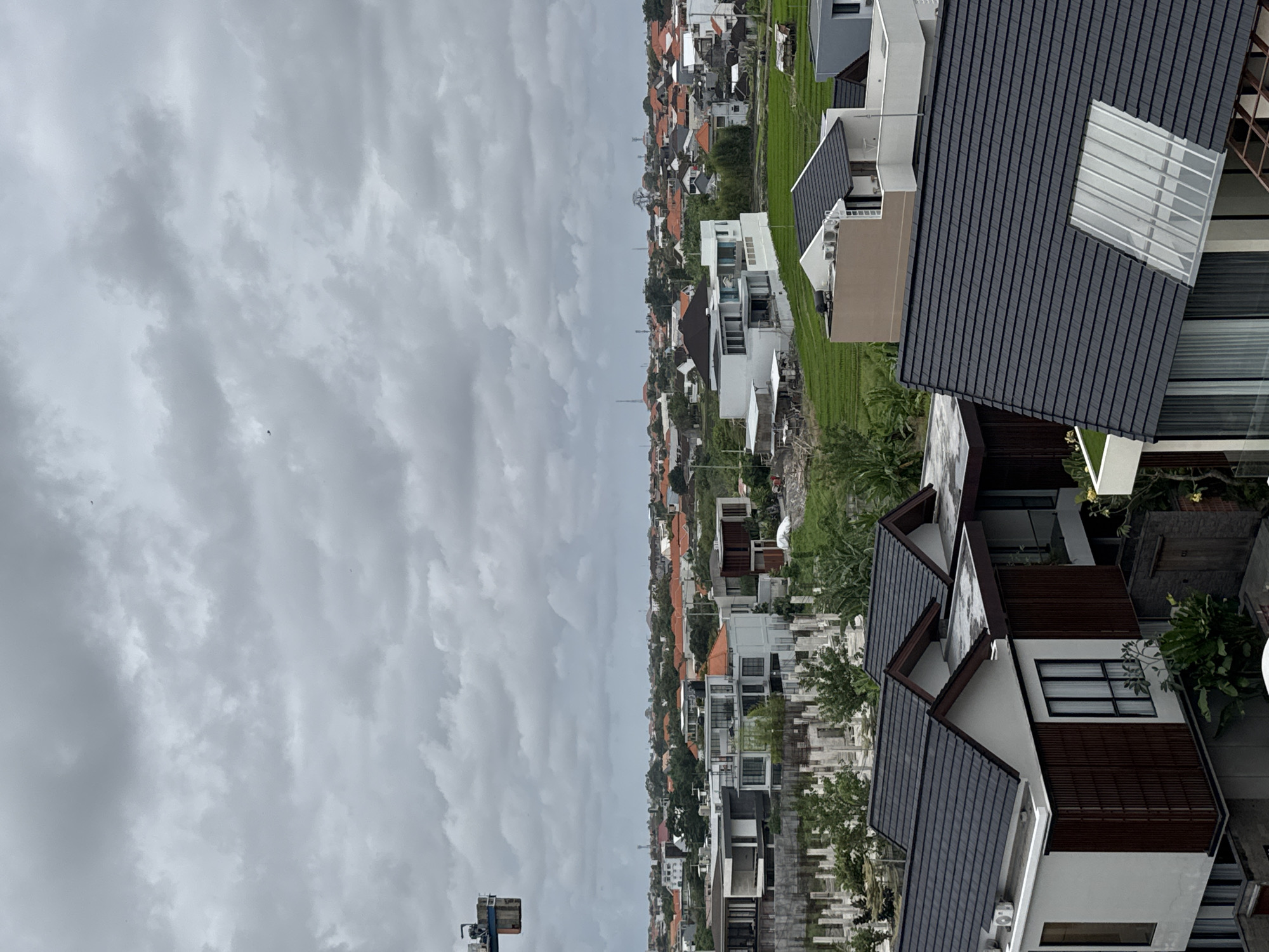 The image depicts an overcast sky above a dense urban and semi-urban landscape in North Kuta, Indonesia. The foreground is dominated by the dark, tiled roofs of modern residential buildings, characterized by sharp angles and large windows. To the mid-ground, a variety of houses with orange tiled roofs are visible, interspersed with lush green vegetation and areas of construction with concrete pillars. A vibrant green rice paddy occupies a significant portion of the mid-ground on the right side of the image, with small structures suggesting agricultural use. In the background, the cityscape stretches out, characterized by a mix of residential buildings, some with white facades and flat roofs, and intermittent patches of trees. Communication towers are visible on the horizon. A construction crane is partially visible in the upper left corner of the image. The overall scene suggests a developing area with a blend of modern architecture, traditional housing, agricultural land, and ongoing construction. There are no people visible in the image.