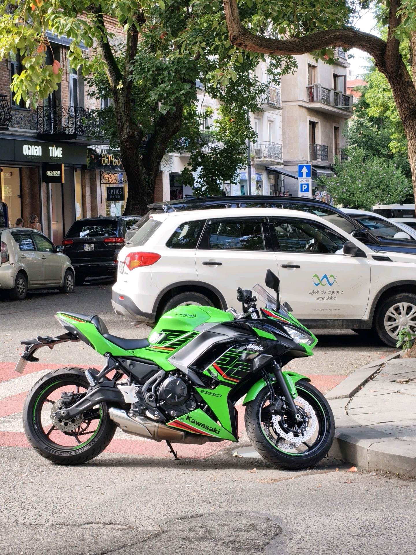 An eye-level, medium outdoor shot captures a vibrant green and black Kawasaki Ninja motorcycle parked on a sidewalk next to a street in Batumi, Georgia. The motorcycle is the primary subject, positioned at a slight angle with its front wheel turned left. The bike features a distinctive green and black paint scheme with red accents and "Ninja" branding on the front fairing. The "Kawasaki" logo is visible on the lower fairing.
Behind the motorcycle, two white SUVs are parked on the street. The one closer to the viewer has Georgian text and logos on its side. A black SUV is parked to the left of the white SUVs. Further to the left, a silver hatchback car is parked.
The background includes a multi-story building with balconies and arched windows, typical of European architecture. Trees with lush green foliage line the street, casting shadows on the ground and partially obscuring the buildings. A street sign with a blue upward arrow and a "P" for parking is visible on a pole. The street itself is paved, with distinct red and white markings of a crosswalk visible beneath the motorcycle. The sidewalk is concrete, showing signs of wear and cracking. The overall scene suggests a bright, sunny day.