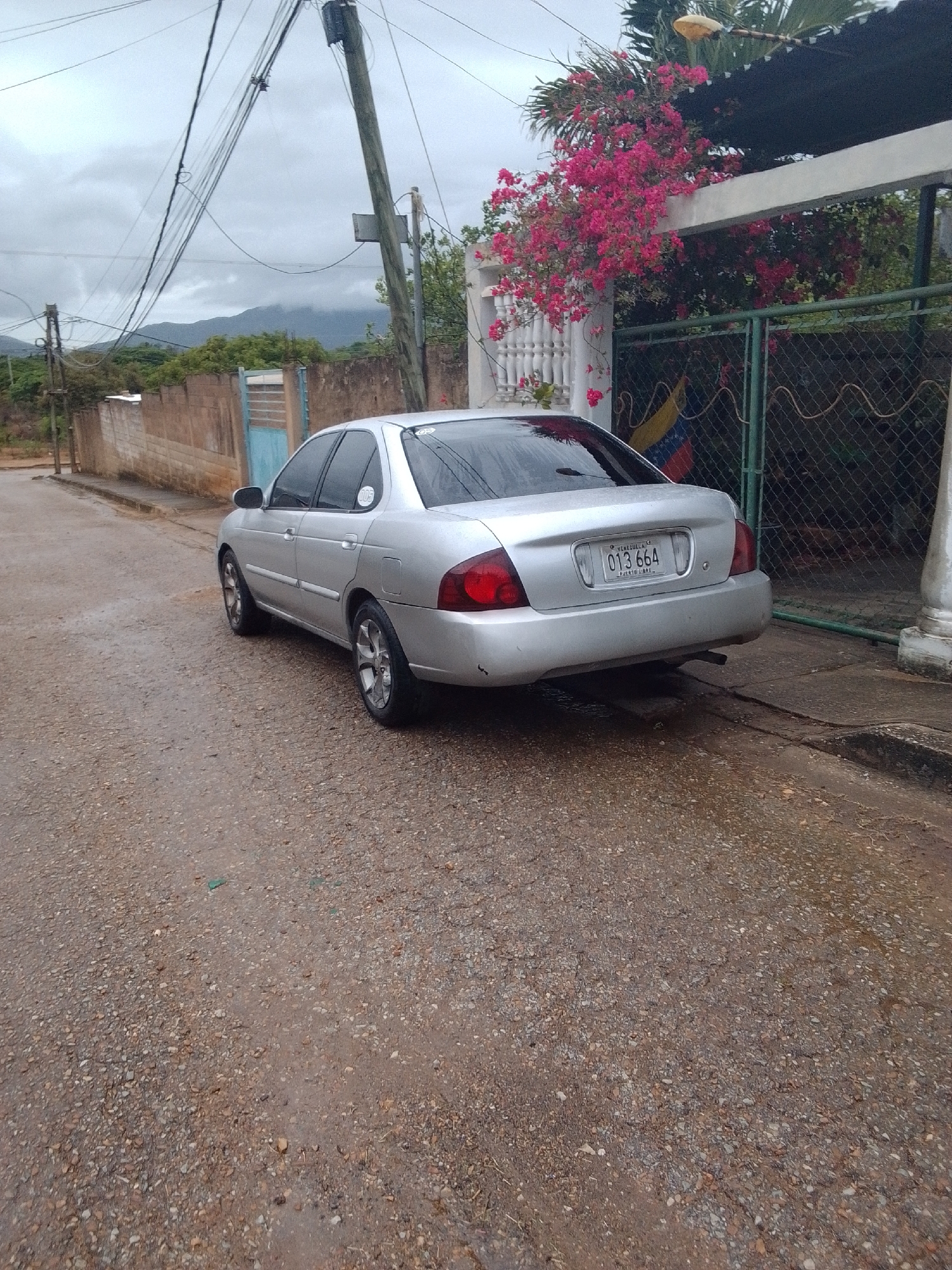 The media file displays a silver sedan parked on a wet, gravelly road in what appears to be a residential area. The car is the primary subject, seen from a rear-three-quarter angle. Its license plate is clearly visible, reading "013 664" with "VENEZUELA" and "PUERTO LIBRE" printed below.
The setting includes a textured concrete wall on the left, partially obscured by lush pink bougainvillea flowers cascading from above. Behind the car, a green chain-link fence with a Venezuelan flag hanging on it can be seen. In the background, there are houses with muted colors, electrical poles with numerous wires crisscrossing, and distant, hazy mountains under an overcast sky.
The overall impression is that of a cloudy day, possibly after rain, given the wet road surface and the overcast conditions. The scene suggests a quiet, possibly rural or suburban setting in La Tagua, Venezuela. There are no people visible in the image, and the car appears stationary, implying no active event is taking place other than the vehicle being parked. Notable details include the vibrant pink flowers contrasting with the muted tones of the surroundings and the prominent Venezuelan flag.
