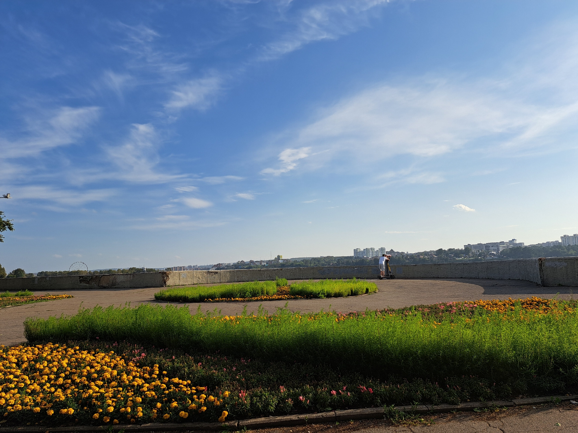 The image shows a park with yellow flowers in the foreground, followed by a patch of green grass and a grey concrete wall. Two people are visible near the wall, one on an electric scooter and the other standing next to them. In the background, a city skyline with buildings and a Ferris wheel can be seen under a blue sky with scattered white clouds. The scene is consistent with the embankment of the Angara River in Irkutsk, Russia.