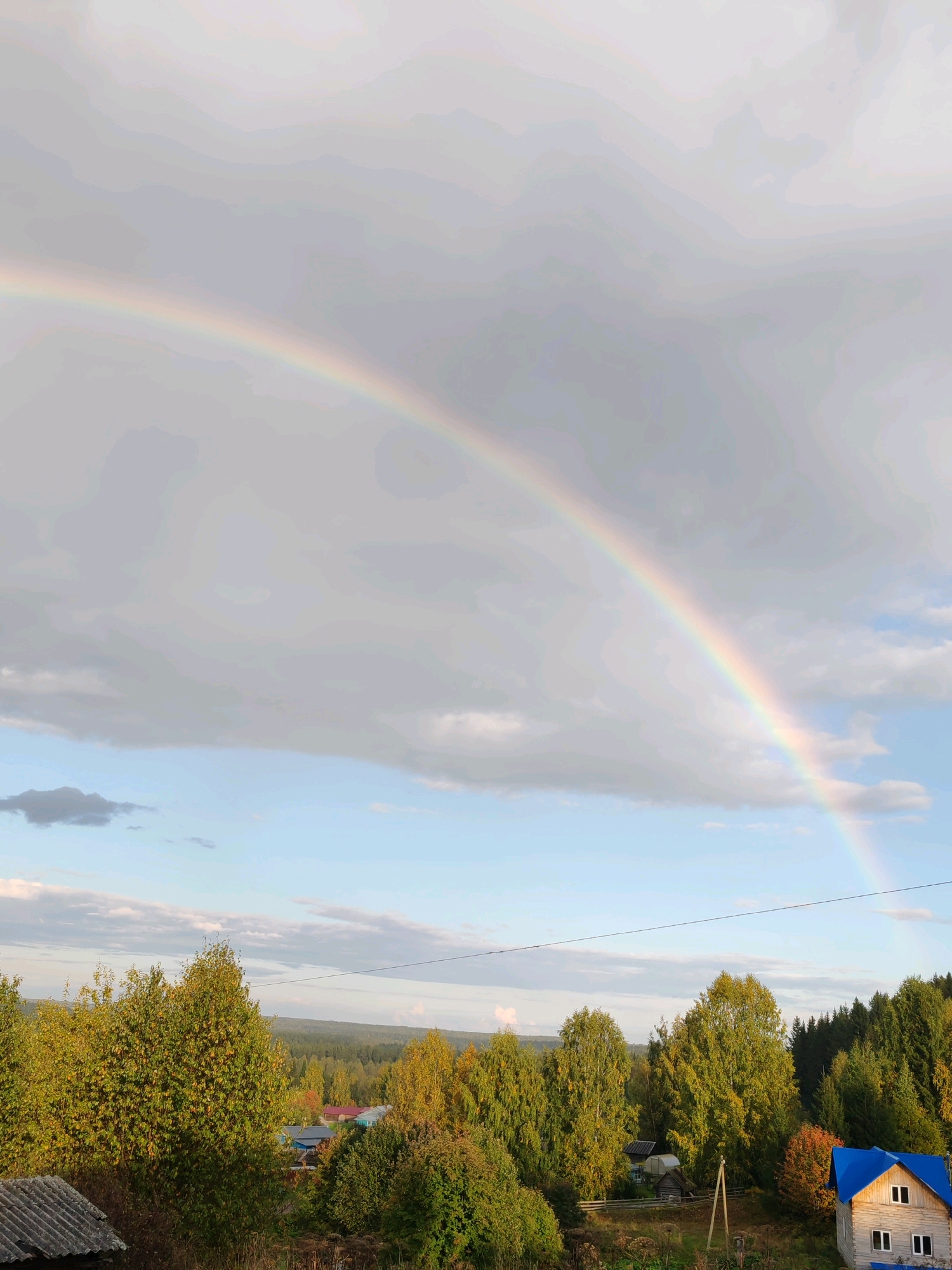 In the city of Chukhlem, Russia, a double rainbow arcs across a grey, cloudy sky, with a sliver of blue visible near the horizon. Below the sky, a dense forest of green and yellow trees stretches across rolling hills. Scattered among the trees are several houses with multi-colored roofs, some appearing to be made of wood. In the foreground, a dilapidated structure with a corrugated roof is visible. The scene is serene and rural, suggesting a post-rainy day atmosphere.