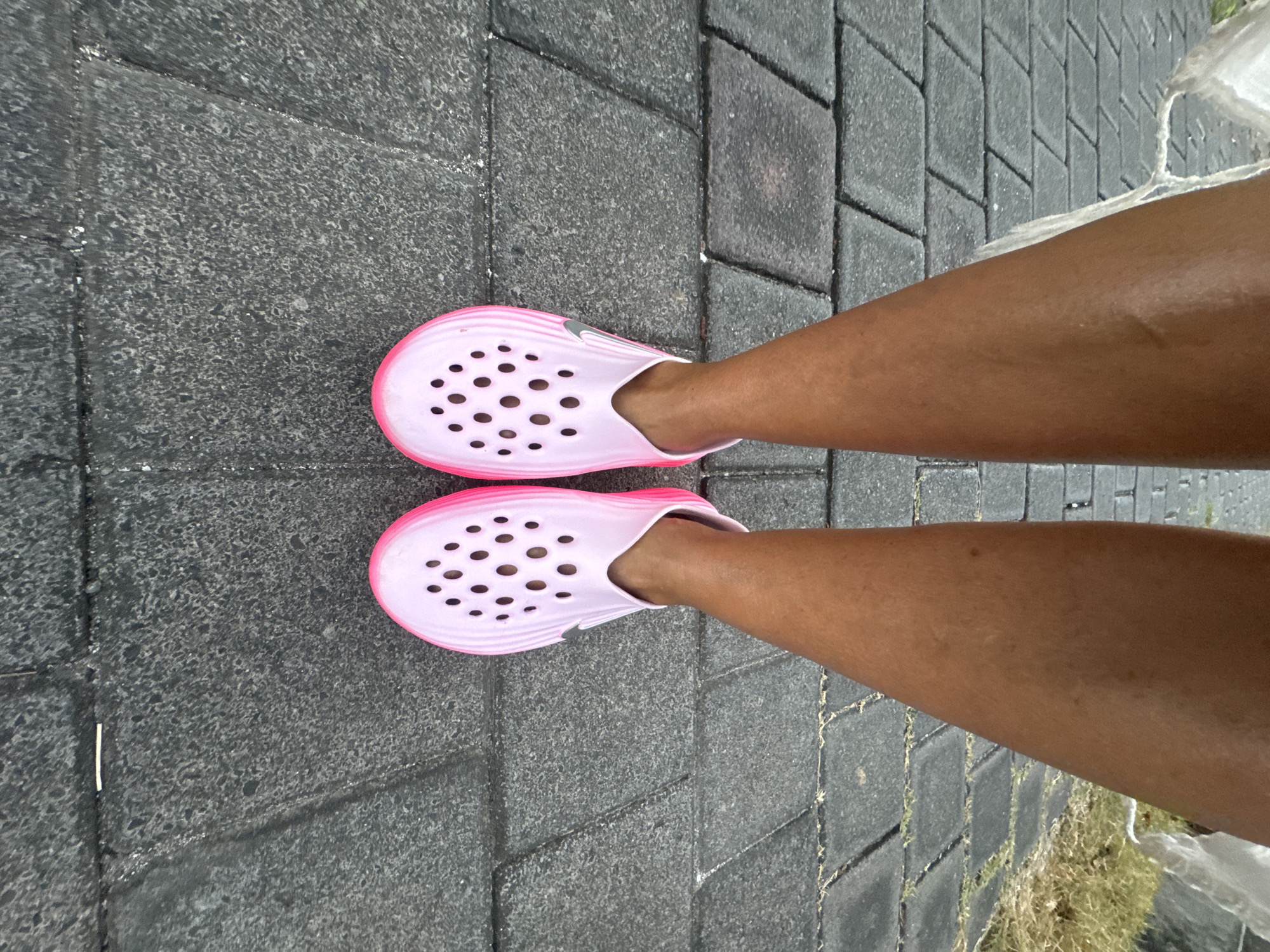 An overhead shot captures a person's legs and feet, clad in light pink/lavender and hot pink clogs, standing on a textured grey paver surface. The perspective looks straight down, with the feet angled slightly outwards.
**Subjects:** The primary subjects are the lower legs and feet of an individual, along with their footwear. The person has a medium to dark skin tone.
**Objects:** The shoes are distinctive clogs, similar to Crocs, featuring numerous circular ventilation holes on the top and sides. They have a bright hot pink sole and trim, with the main body of the shoe being a lighter pink or lavender hue. A small silver Nike swoosh logo is visible on the outer side of each shoe, just above the hot pink trim. The ground is paved with rectangular, interlocking grey concrete pavers, which show some texture and varying shades of grey, indicating possible wear or moisture.
**Scene/Setting:** The scene is an outdoor setting, likely a pathway, sidewalk, or courtyard in Kuta, Indonesia, given the context. The paving stones suggest a pedestrian area.
**Activity or Event:** The person appears to be standing or possibly walking, with their feet planted on the ground. The image captures a moment of rest or transition.
**Notable Details:** The surface of the pavers appears slightly damp or has water droplets on it, suggesting recent rain or a damp environment. Similarly, the skin on the person's legs has a subtle sheen, which could be from moisture or sweat, consistent with a humid climate. Some sparse green vegetation, possibly grass or weeds, can be seen at the right edge of the paved area, suggesting proximity to natural ground. The lighting indicates it is daytime.
**Visible Text:** A silver Nike swoosh logo is visible on the outer side of each pink clog.