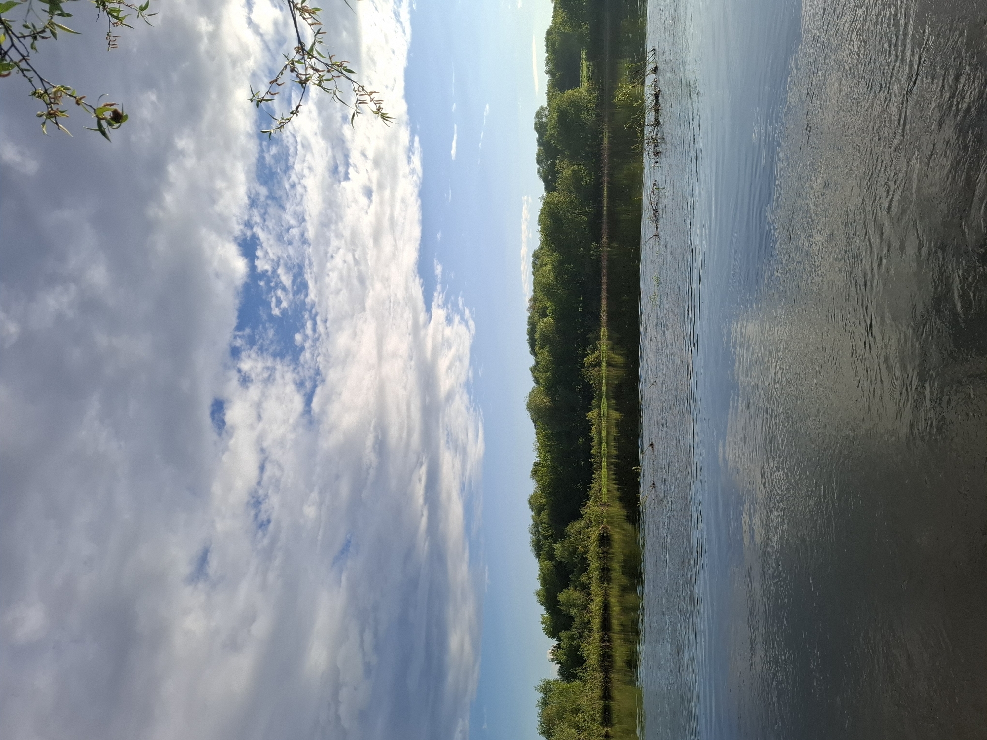 The image shows a view of a body of water, likely a lake or river, with trees lining the opposite bank. The sky is visible with clouds and blue patches. The image appears to be taken in Дерезовка, Russia. There are no people visible in the image. The scene appears to be calm and still.