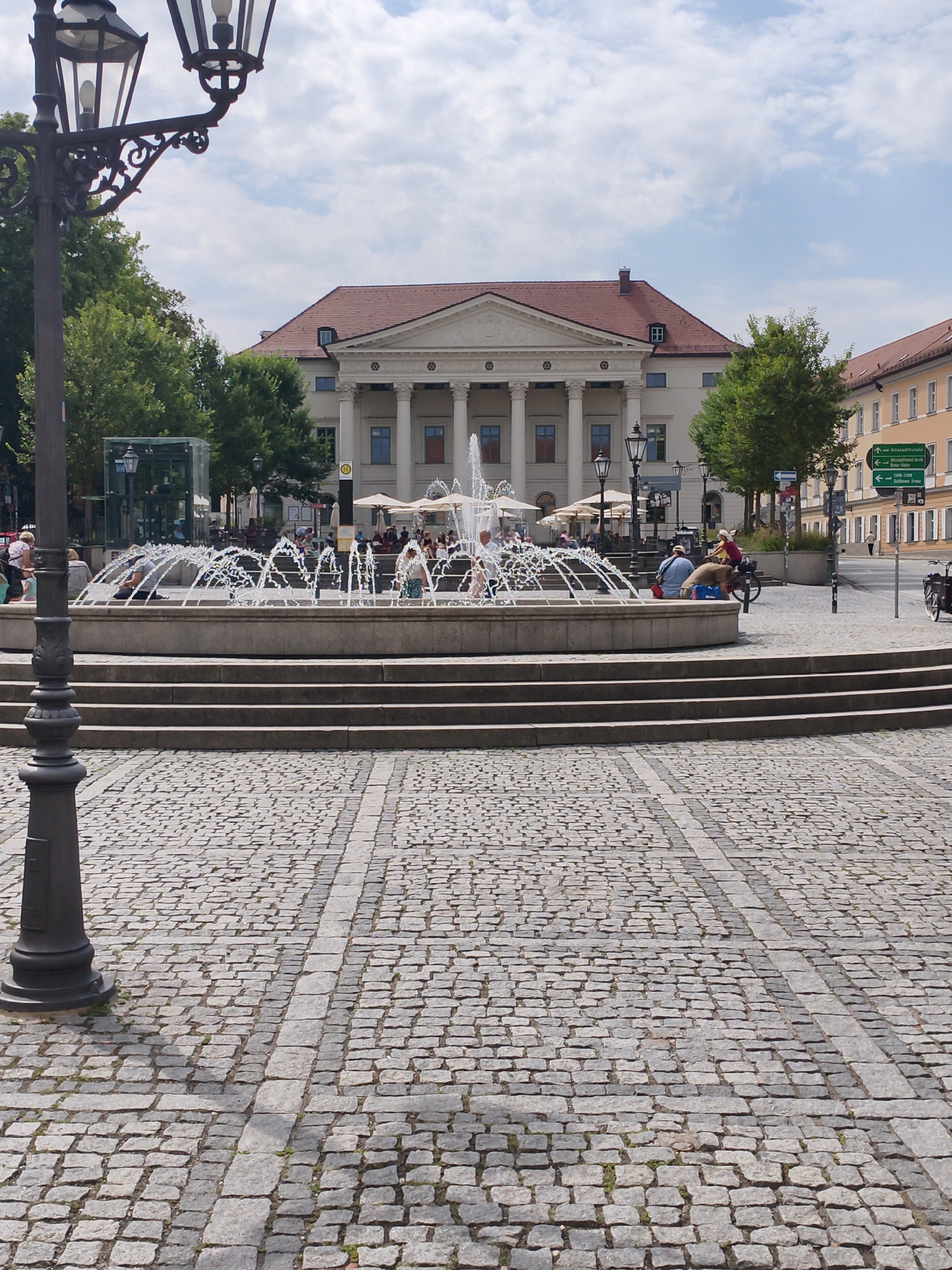 In Regensburg, Germany, on a sunny day, a fountain is spraying water in a public square paved with cobblestones. A classical building with columns stands in the background, and a lamppost is visible in the foreground. People are seen sitting on the fountain's edge, interacting with the water, and relaxing. Several individuals are enjoying the fountain's spray, while others are seated nearby. The scene depicts a lively, community-oriented atmosphere within the historic cityscape of Regensburg.