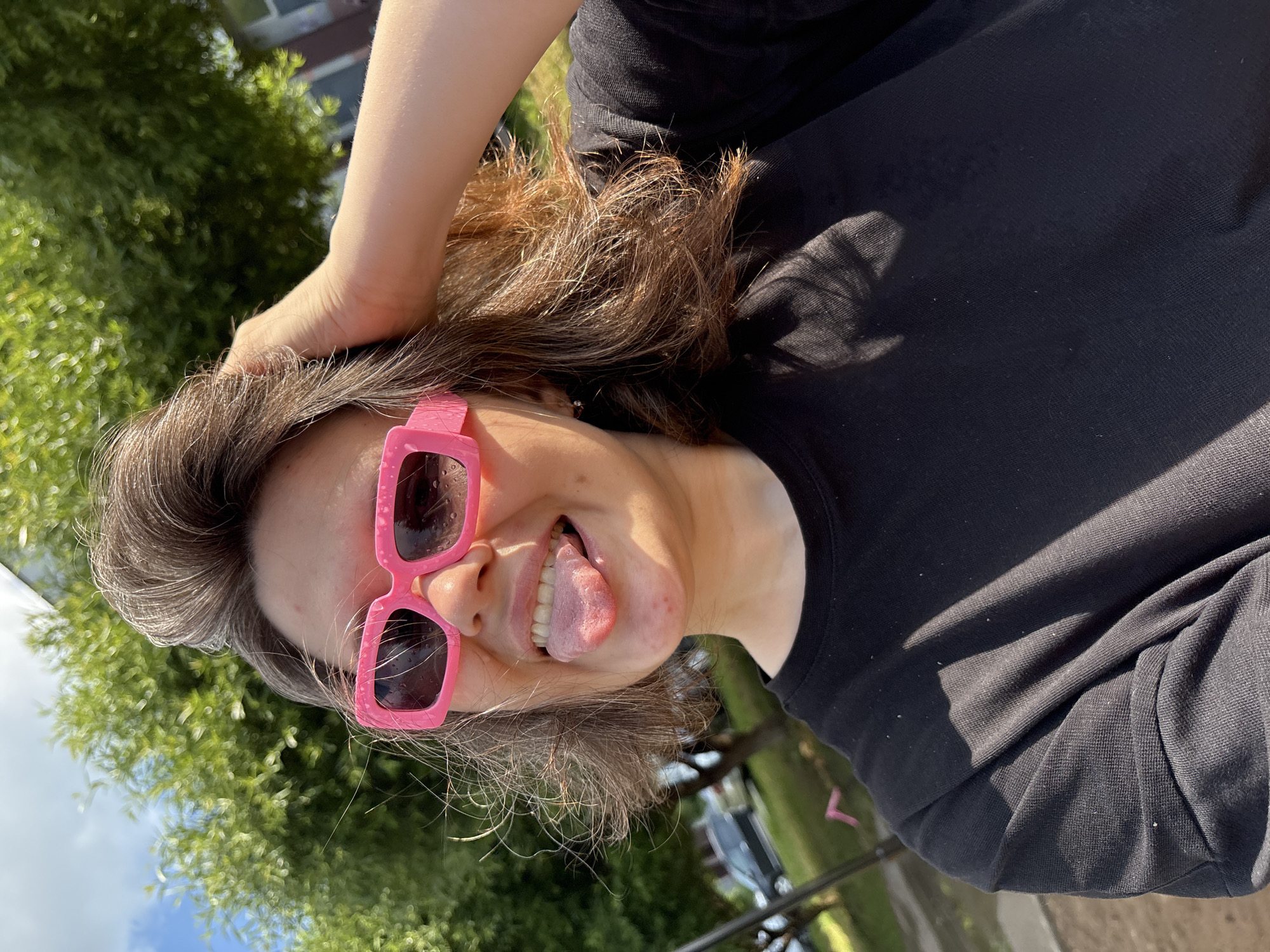 In a medium shot, a young woman with brown hair, wearing bright pink sunglasses and a black t-shirt, sticks her tongue out at the camera. Her hand is raised and resting on her head. The background features green trees and a bright sky, suggesting an outdoor setting. The image appears to be a selfie taken on a sunny day. The presence of trees and buildings in the background, along with what looks like a parking area with a car, indicates an urban or suburban environment. The light suggests it is daytime, likely afternoon due to the angle of the sun casting shadows. The overall impression is one of a casual, lighthearted moment.