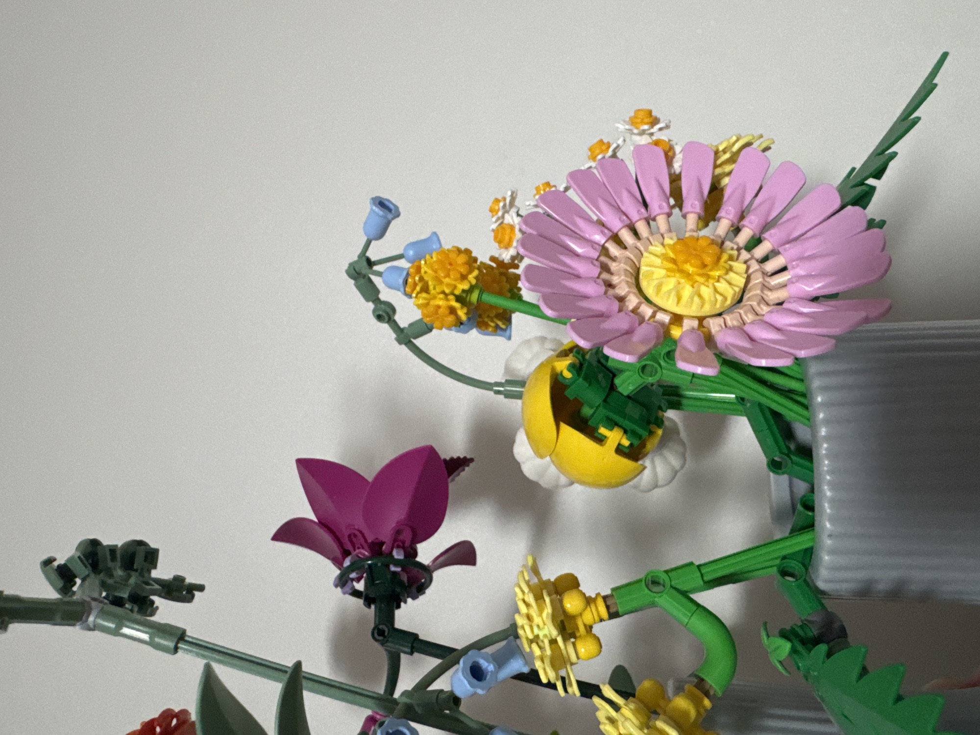 This image shows a close-up of a Lego flower arrangement in a grey vase. The arrangement includes several types of flowers constructed from Lego bricks in various colors, including pink, yellow, white, orange, and blue. The background is a plain, light-colored wall, suggesting an indoor setting. There is no indication of the location being Kyiv, Ukraine. The scene is static and focuses solely on the Lego display.