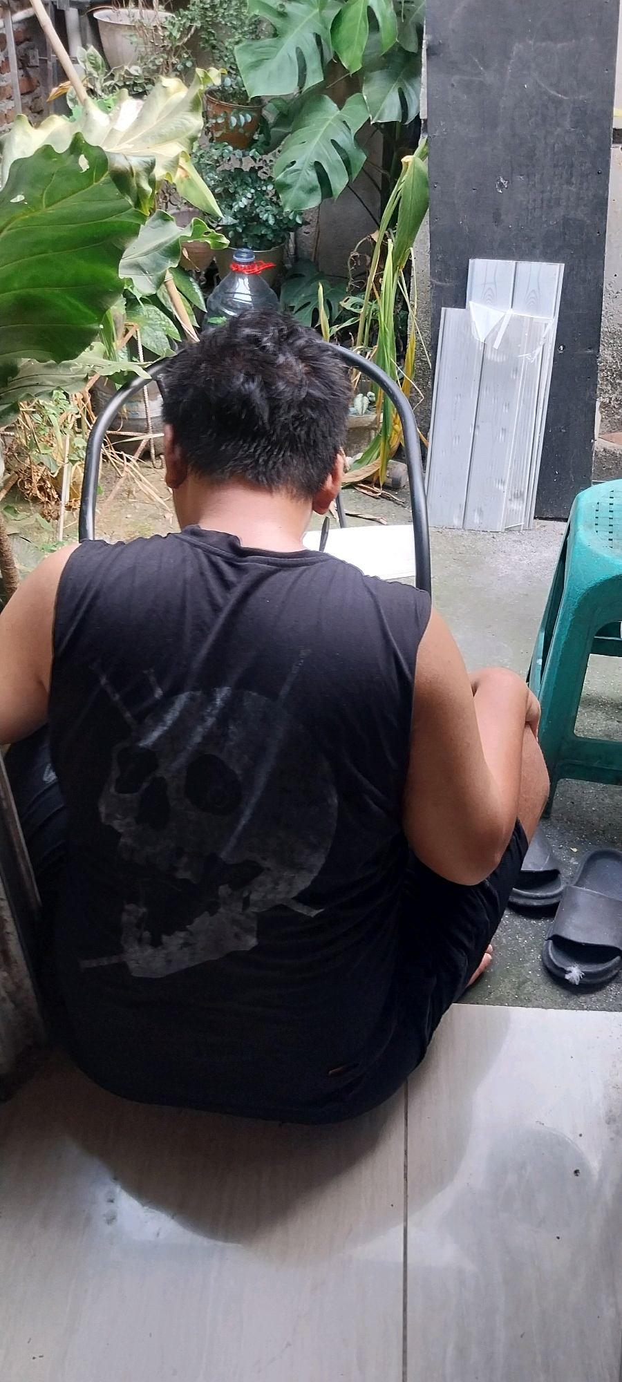 In Bojong Nangka, Indonesia, a person sits facing away from the camera, on a metal chair, in an outdoor area. They are wearing a black sleeveless shirt with a skull graphic printed on the back and black shorts. The person is seated on a tiled floor, with a pair of black sandals visible to their right. Behind them, a variety of potted plants and foliage are visible, suggesting a garden or backyard setting. The environment appears to be somewhat cluttered, with some plastic sheeting or panels leaning against a dark wall in the background. A green plastic stool is also present in the scene. The lighting suggests it is daytime.
