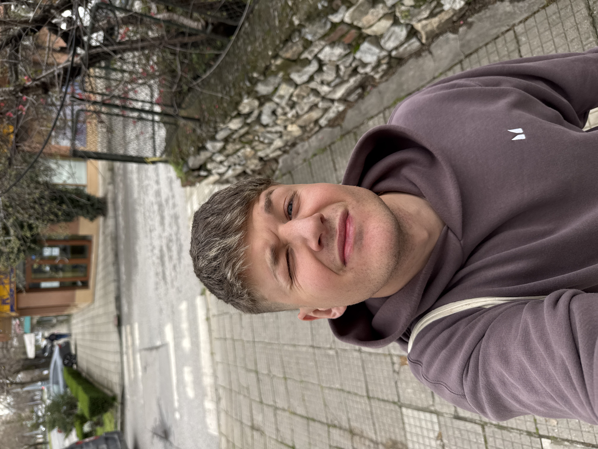 A light-skinned male with short grey hair is taking a selfie while standing on a sidewalk paved with square, light-grey tiles. The man is wearing a mauve-colored hooded sweatshirt with a small white logo on the upper left chest. He is winking his left eye. Behind him is a street, on the other side of which is a building with a brown-framed window. There are several cars parked on the street. A stone wall runs alongside the sidewalk to the man's right. Bare branches of trees are visible above the building and the stone wall. The location is Plovdiv, Bulgaria.