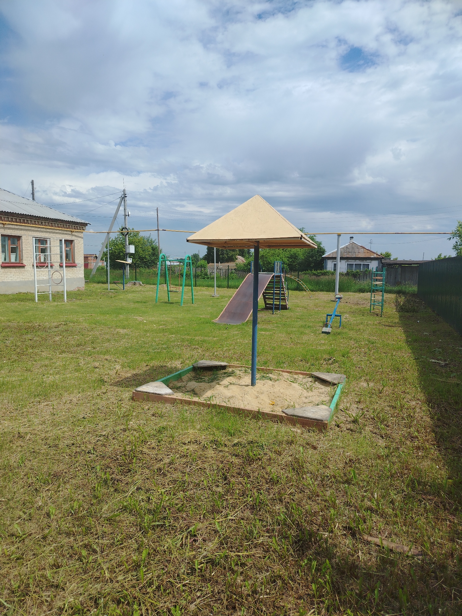 This is an exterior, eye-level, medium shot of a playground in the city of Klyuchi, Russia, on a cloudy day. The playground features a sandbox with a triangular, yellow umbrella, a metal slide with blue steps, a blue seesaw, a green swing set, and a ladder climbing structure. In the background, there is a brick building, a house with a tiled roof, and a green fence. The ground is covered in green grass with some patches of dry, matted grass. There are no people present in the image.