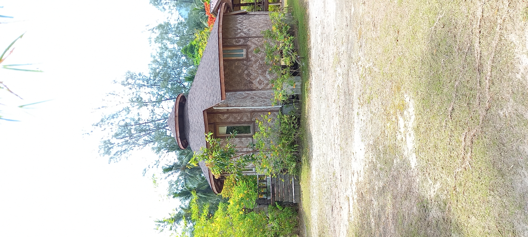 The image shows a small, single-story hut in Baan Chaloklum, Thailand. The hut is constructed of woven materials and features a distinctive roof design with a conical top. It has at least two windows visible on its facade. The hut is surrounded by greenery, including trees, bushes, and flowering plants. A set of stairs leads to the entrance of the hut. Next to the hut is a dirt path. The sky is partly visible, but mostly obscured by trees.