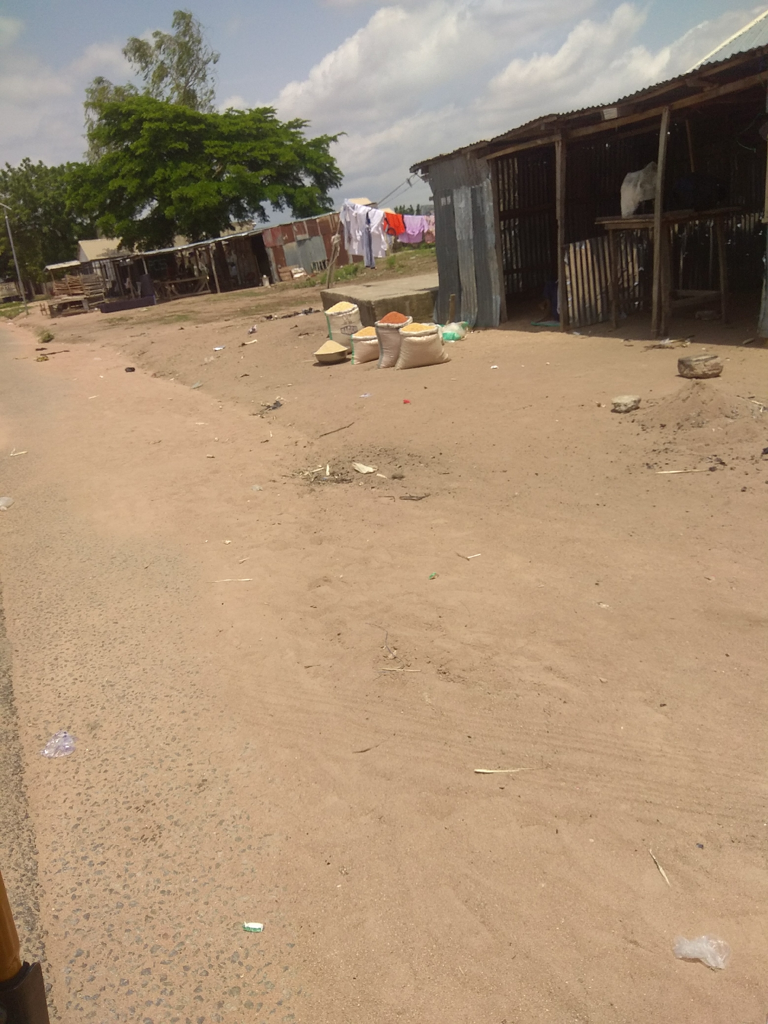 The image depicts a street scene in Jalingo, Nigeria. A market stall constructed with corrugated metal and wood stands on the right side of the frame. Several sacks, likely containing grains or spices, are placed in front of the stall. Laundry, including shirts and trousers, is hung on a line to the left of the stall. The ground is predominantly dirt and sand, with some scattered debris. A paved road or pathway runs along the left side of the image. In the background, a large green tree is visible, and the sky is partly cloudy. There are no people or animals clearly visible in the scene.
