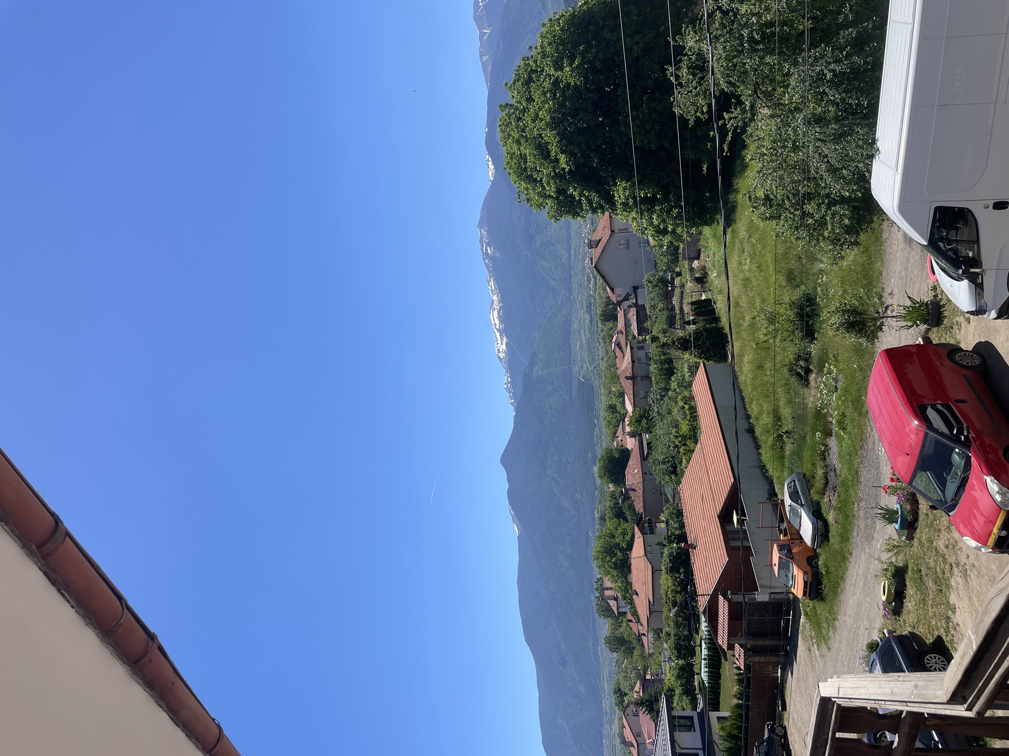 The image depicts a view from an elevated position, possibly a balcony, overlooking a residential area near the city of Yahinovo, Bulgaria. In the foreground, a partial view of a building roof and a wooden railing are visible. The middle ground contains several houses with red-tiled roofs interspersed with trees and vegetation. Parked vehicles, including a red car, a white van, and a silver car, are visible on a pathway. In the background, a mountain range with some snow-capped peaks forms the horizon against a clear blue sky. A contrail from a jet plane can be seen in the upper left portion of the sky. Power lines run through the center of the image.