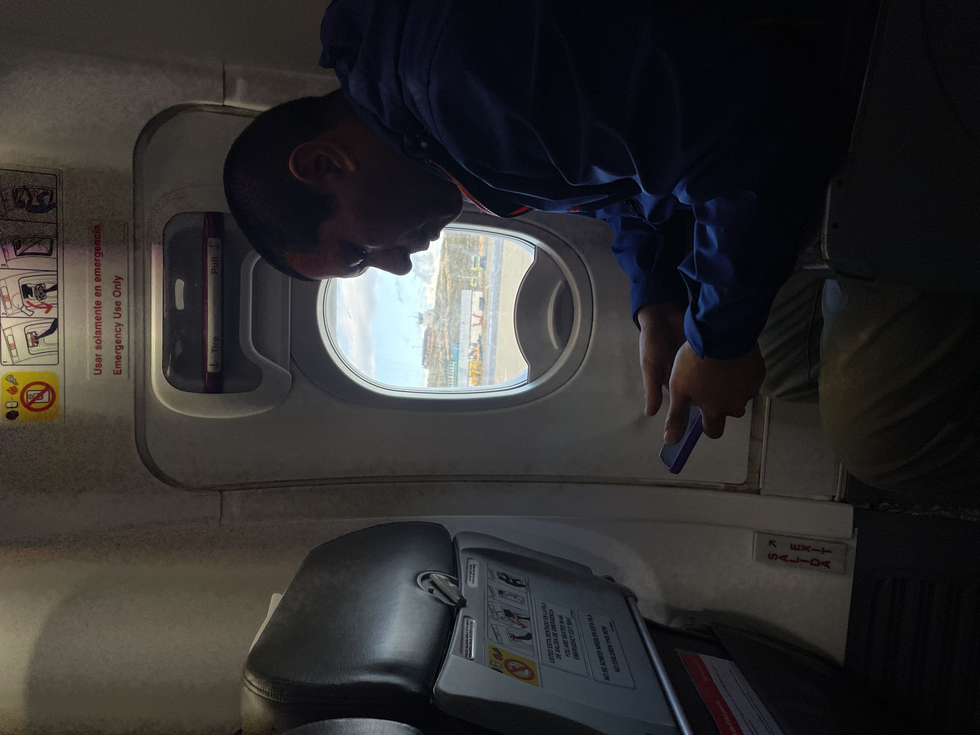 The image captures an airplane interior, likely at an airport in Ushuaia, Argentina, viewed from a slightly low angle across an aisle. A person wearing a dark blue jacket leans forward, looking out a circular airplane window. Their face is mostly in shadow, but their right hand is visible holding a purple-cased smartphone, suggesting they are observing or photographing the view.
Through the window, an airport tarmac is visible, featuring buildings, ground vehicles, and what appears to be a cloudy or overcast sky, consistent with a daylight scene. The airplane's emergency exit door dominates the left side of the frame, displaying prominent bilingual labels. Text on the door reads "Usar solamente en emergencia / Emergency Use Only" and "Pull / Tire" with directional arrows.
In the foreground, a dark-colored airplane seat with a partially lowered seatback tray table is visible. The tray table displays detailed safety instructions and bilingual warnings: "USTED ESTA SENTADO EN LA FILA DE SALIDA DE EMERGENCIA / YOU ARE SEATED IN AN EMERGENCY EXIT SEAT" and "NO SE ADMITE NIÑOS EN ESTA FILA / NO CHILDREN THIS ROW". Near the bottom right, an "EXIT / SALIDA" sign is visible on the floor or lower wall. The interior lighting is subdued, creating a contrast with the brighter view outside the window. The provided image itself is rotated 90 degrees clockwise.