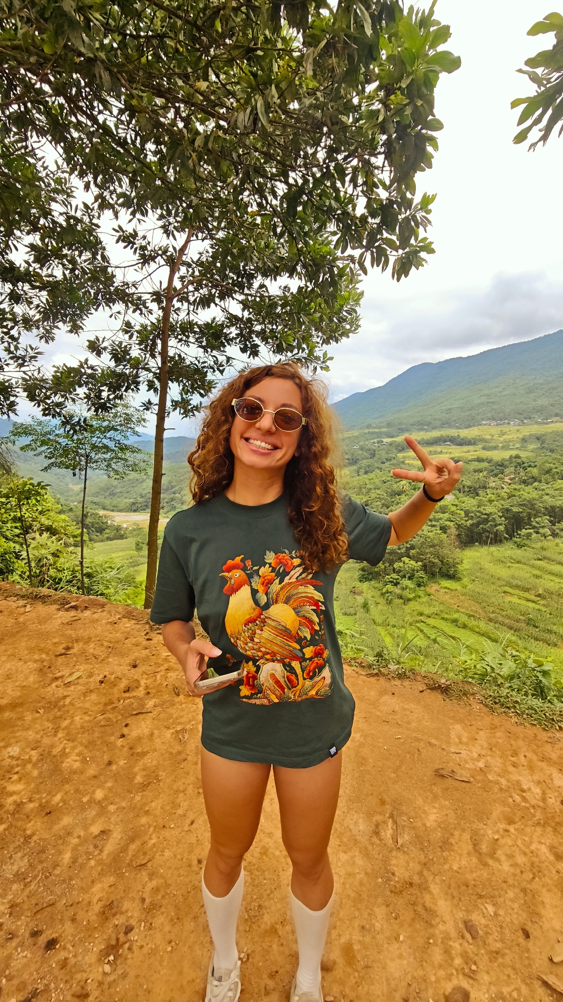 A young woman stands on a dirt path, smiling at the camera. She has curly brown hair and wears round sunglasses, a dark green t-shirt with a rooster and floral graphic, white socks, and sneakers. Her right hand is raised, forming a peace sign, and her left hand holds a smartphone. Behind her, a lush green valley stretches out, with terraced rice fields, a small village, and distant mountains under a cloudy sky. The location is Pù Luông Commune, Vietnam.