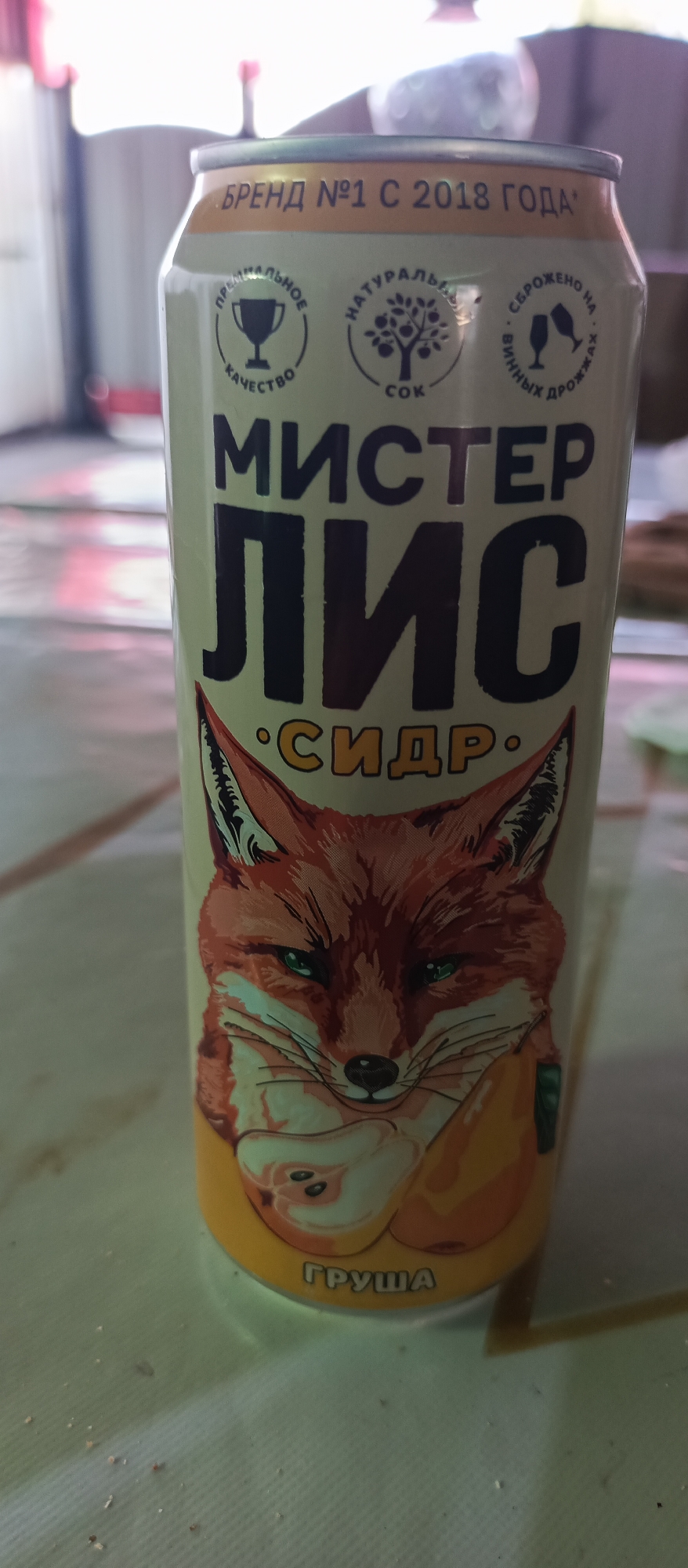 The image shows a close-up of a can of "Mister Lis" pear cider. The can features a cartoon illustration of a fox wearing a green scarf and holding a pear. The text on the can is in Russian and includes the brand name "Mister Lis," the product type "Cider," and the flavor "Pear." There are also several seals on the can, indicating "Premium Quality," "Natural," and "Fermented on wine yeast." The can is placed on a table with a patterned tablecloth. The background is blurred, but it appears to be an indoor setting.