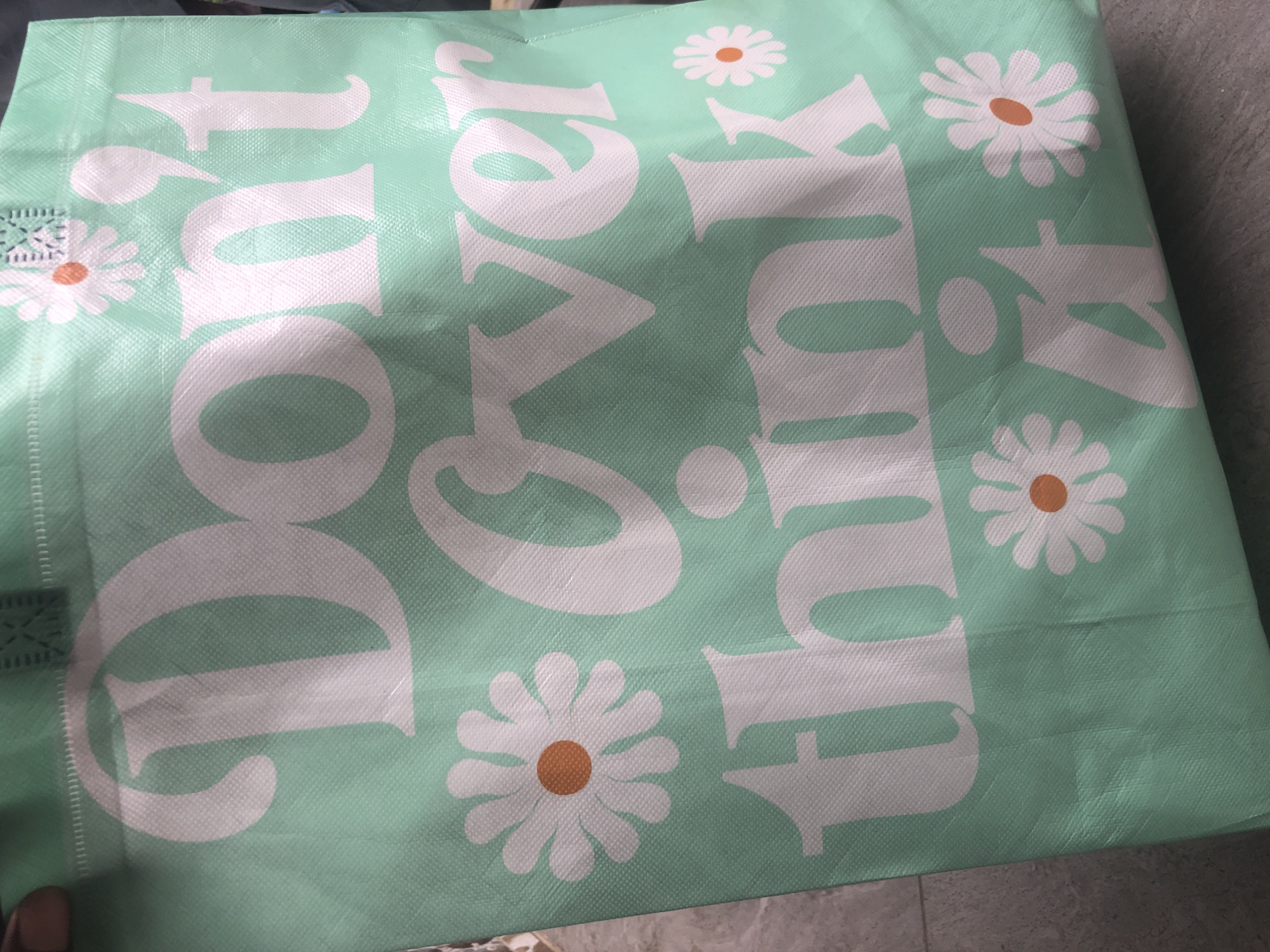 The image shows a close-up of a light green reusable shopping bag with white text and daisy graphics. The text reads "Don't Over think it". The bag is made of a textured material. The background appears to be a concrete surface. There is no information in the image that indicates a specific location in Ado, Nigeria.