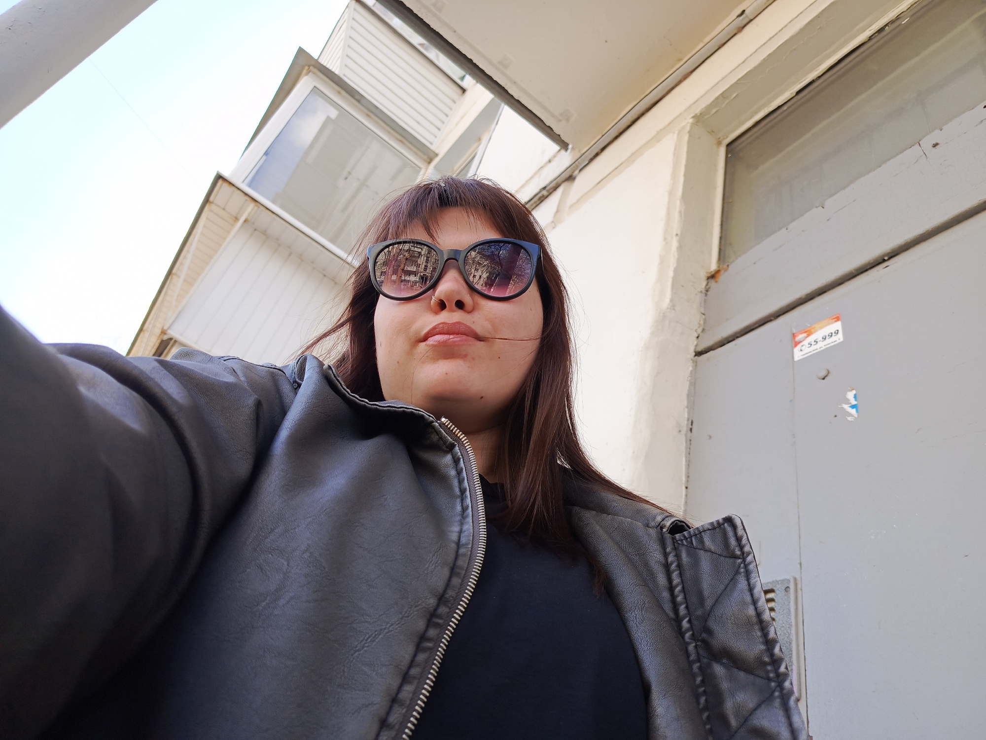 A low-angle selfie captures a young person, likely female, with medium-length dark brown hair and bangs, wearing black-framed sunglasses with reflective pinkish-purple lenses. A nose piercing is visible. She is dressed in a dark, possibly black or dark gray, leather-look jacket featuring a prominent silver zipper, and a dark shirt underneath.
The bright daylight indicates clear weather, with a light blue sky visible in the upper left portion of the image. The reflections in the sunglasses clearly show bare tree branches and densely packed buildings, suggesting an urban or suburban outdoor setting, likely in Chaikovsky, Russia.
The background consists of the exterior of a building, likely a residential block, characterized by light-colored (white or light gray) walls, windows, and architectural elements that appear to be balconies or extensions. To the right, a light gray door or wall features a small white and orange sticker. The most prominent text on this sticker is "55-999," with other less clear text above it. The person's expression is neutral, largely obscured by the sunglasses. The image captures an ordinary moment in a residential environment.