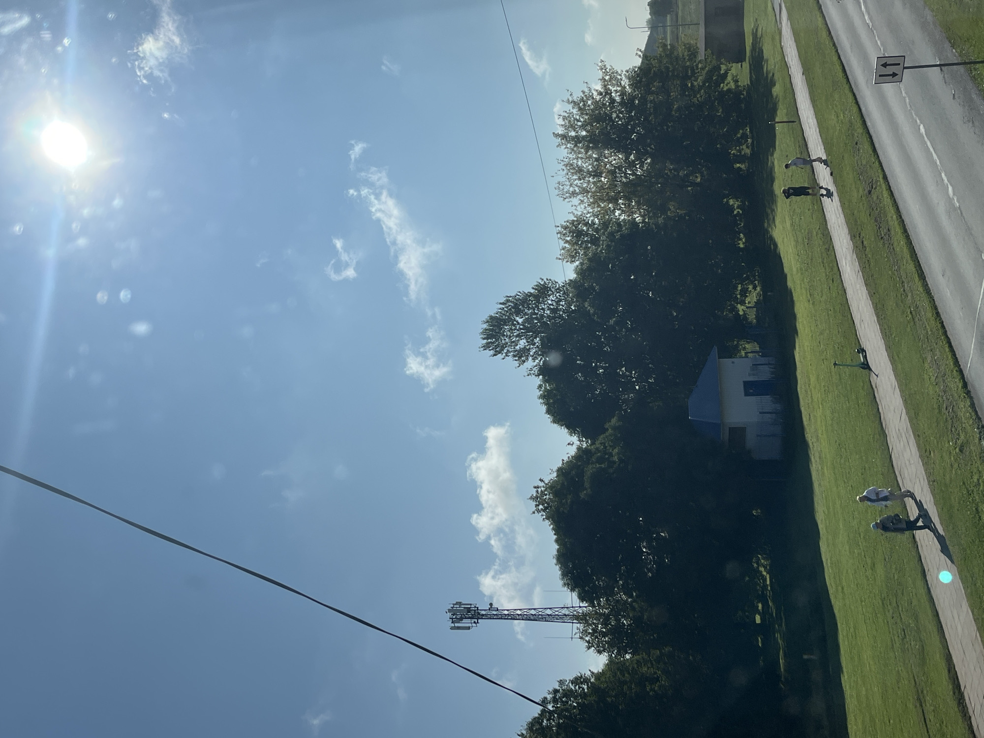 The image depicts an outdoor scene under a bright, sunny sky with scattered clouds, possibly in Kohtla-Järve linn, Estonia. A prominent power line or cable diagonally crosses the upper left portion of the frame. On the left, a tall telecommunications tower stands against the blue sky, surrounded by lush green trees. In the mid-ground, a small white building with a blue roof is visible. A paved walkway runs horizontally across the image, bordered by a grassy area on the left and a road on the right. Several individuals are present: two people are walking on the walkway in the foreground, appearing to be engaged in conversation or walking together. Further down the walkway, two more individuals are seen on what appear to be electric scooters. A green electric scooter is also parked on the side of the walkway. The road surface is visible on the right, with a traffic sign indicating two-way traffic. The overall environment appears to be a park or recreational area adjacent to a road.