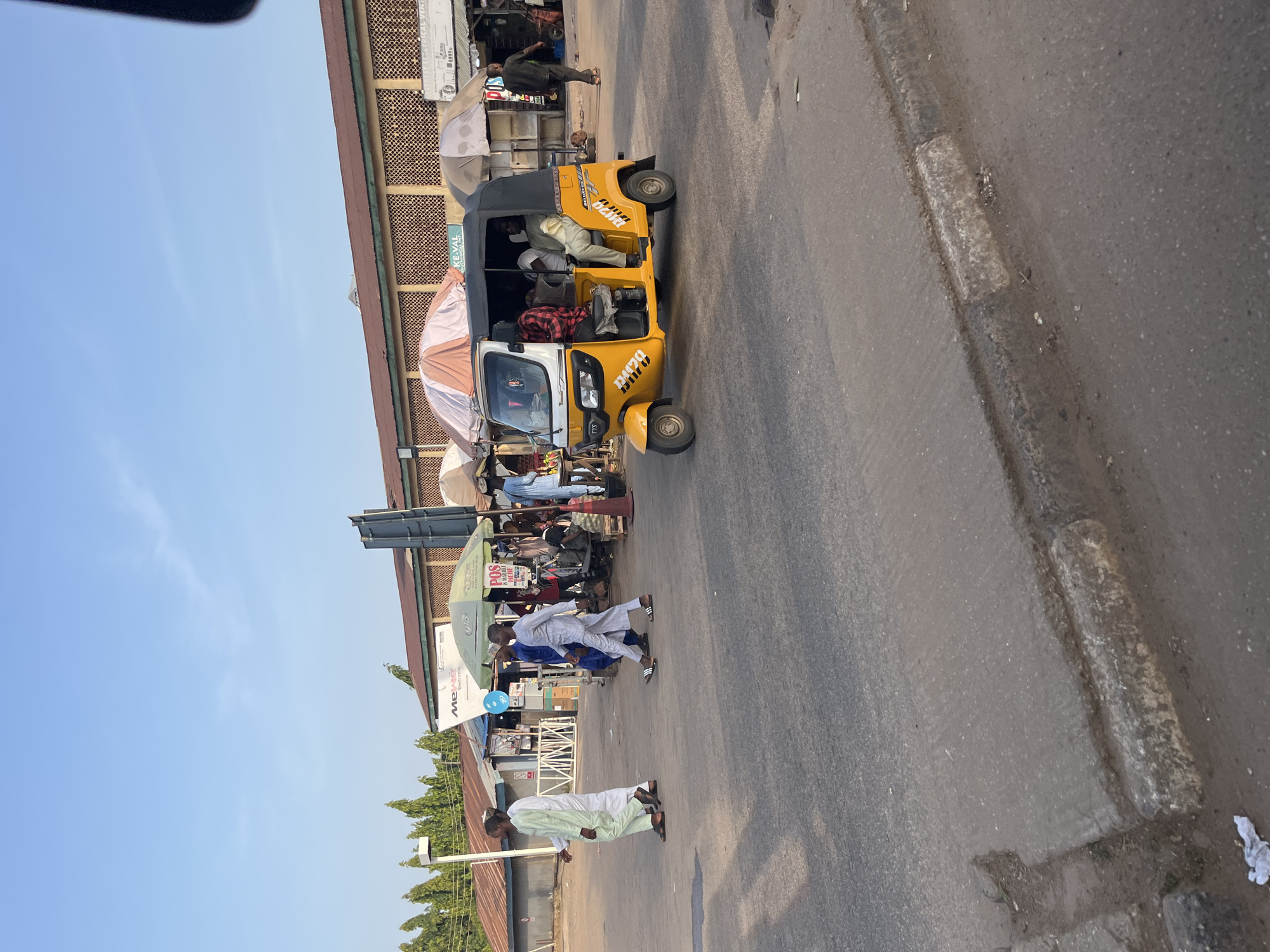 The image captures a street scene in an outdoor market environment, likely in a city in Nigeria. In the foreground, a yellow auto-rickshaw, also known as a 'keke napep', is the central focus. It has a black canopy and is occupied by several people. One person, wearing a checkered shirt, is in the driver's seat, while others are passengers. The auto-rickshaw has prominent markings, including the number "R179" on the side and the brand name "TVS".
Several individuals are walking on the paved road and sidewalk. Two men in traditional white and light green attire are walking from left to right. Another man in a striped white and grey outfit walks in the same direction, closer to the auto-rickshaw. In the background, a row of market stalls or shops with brown roofs is visible. Some stalls are shaded by umbrellas, one with orange and white stripes and another white one. Signs for businesses, including "Mejabo Electronics" and "POS" are present. The general environment appears to be dusty and sunlit, suggesting a warm climate. The sky is a clear, light blue with a few faint clouds. The scene depicts a typical day in a bustling marketplace, with people engaged in commerce and transportation.