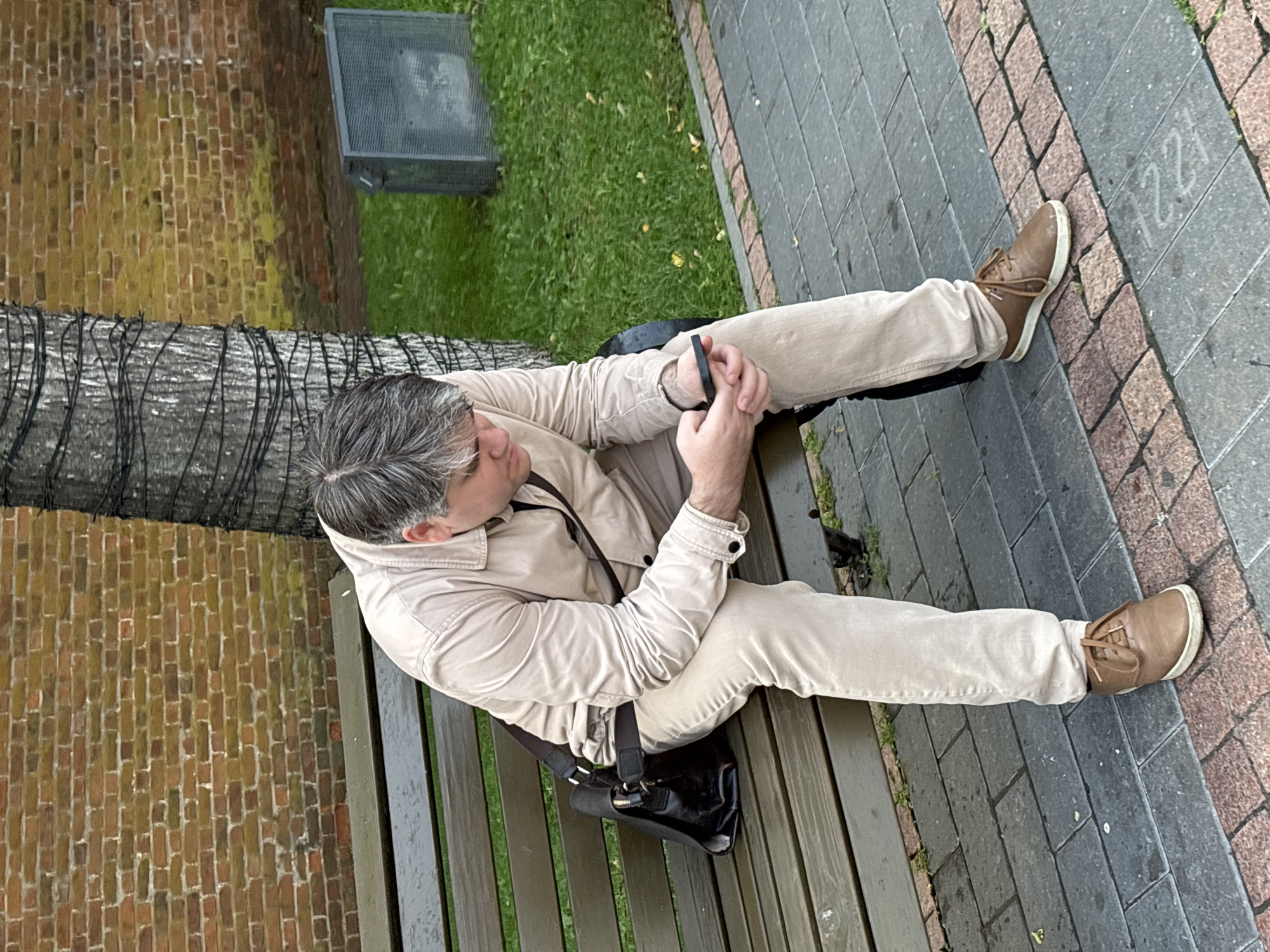 A man with graying hair is sitting on a wooden bench, looking down at his phone. He is wearing a light beige jacket and pants, and brown shoes. A black bag is slung over his shoulder and rests on the bench next to him. The bench is in a park-like setting with a brick wall in the background and a tree with string lights wrapped around it. The ground is paved with interlocking bricks. The image is taken from a slightly elevated angle. The man is looking down at his phone, his left hand holding it and his right hand resting on his leg. The scene appears to be outdoors, possibly in a public area.