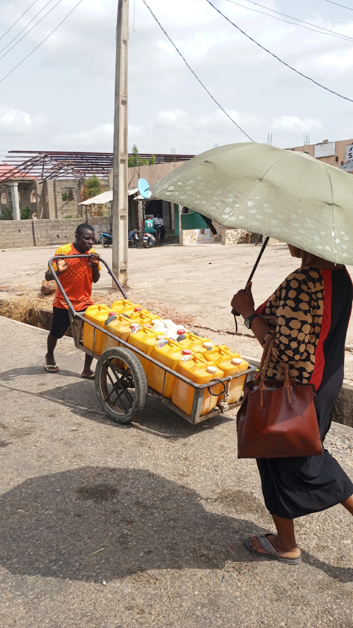 In Zango-ndaji, Nigeria, a man in an orange shirt pulls a two-wheeled metal cart filled with approximately 20 yellow plastic containers. A woman with a brown handbag and an umbrella walks beside the cart. The location is an urban street with buildings visible in the background, including one under construction. Utility poles and power lines are present. Two people sit on the stoop of a building behind the man pulling the cart, and two people are riding a motor bike.